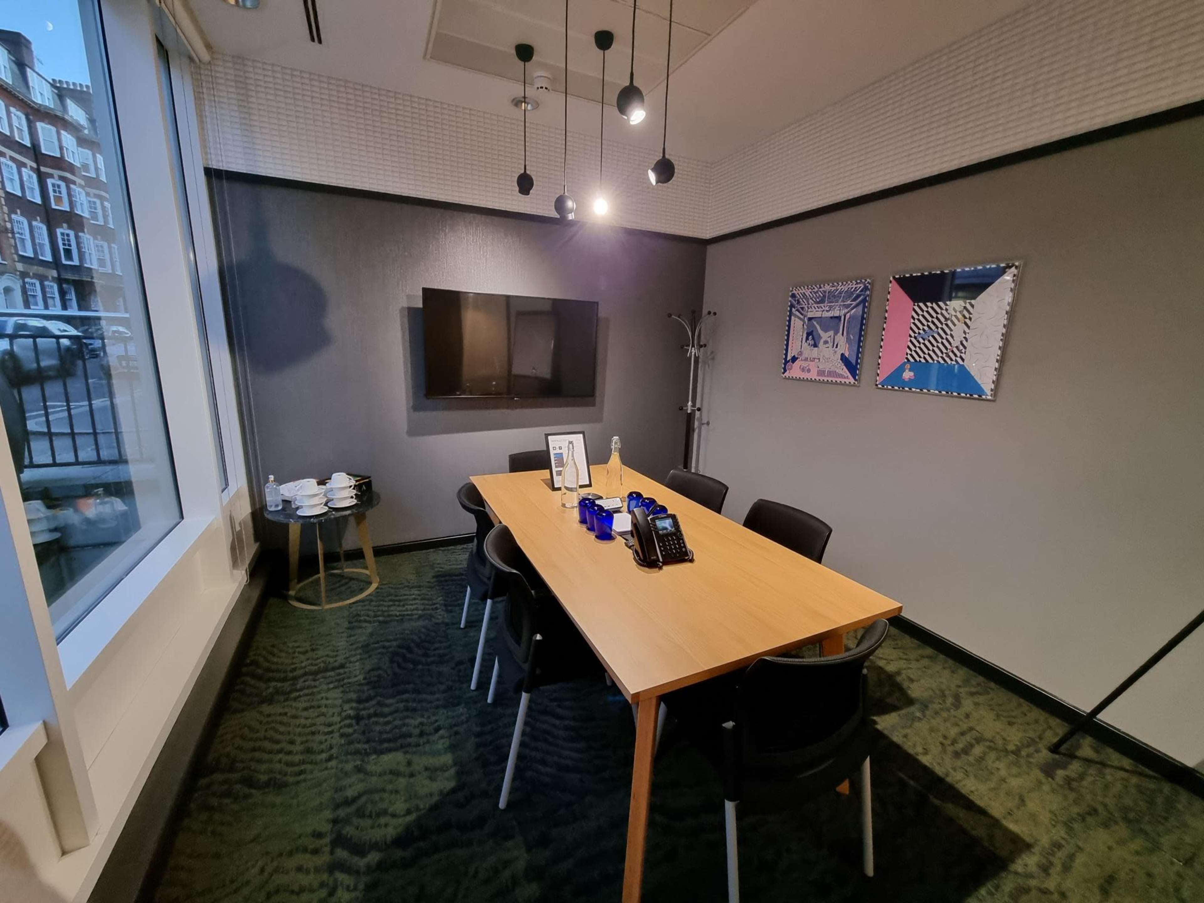 The image shows a conference room with a wooden table, black chairs, a wall-mounted television, and framed artwork.