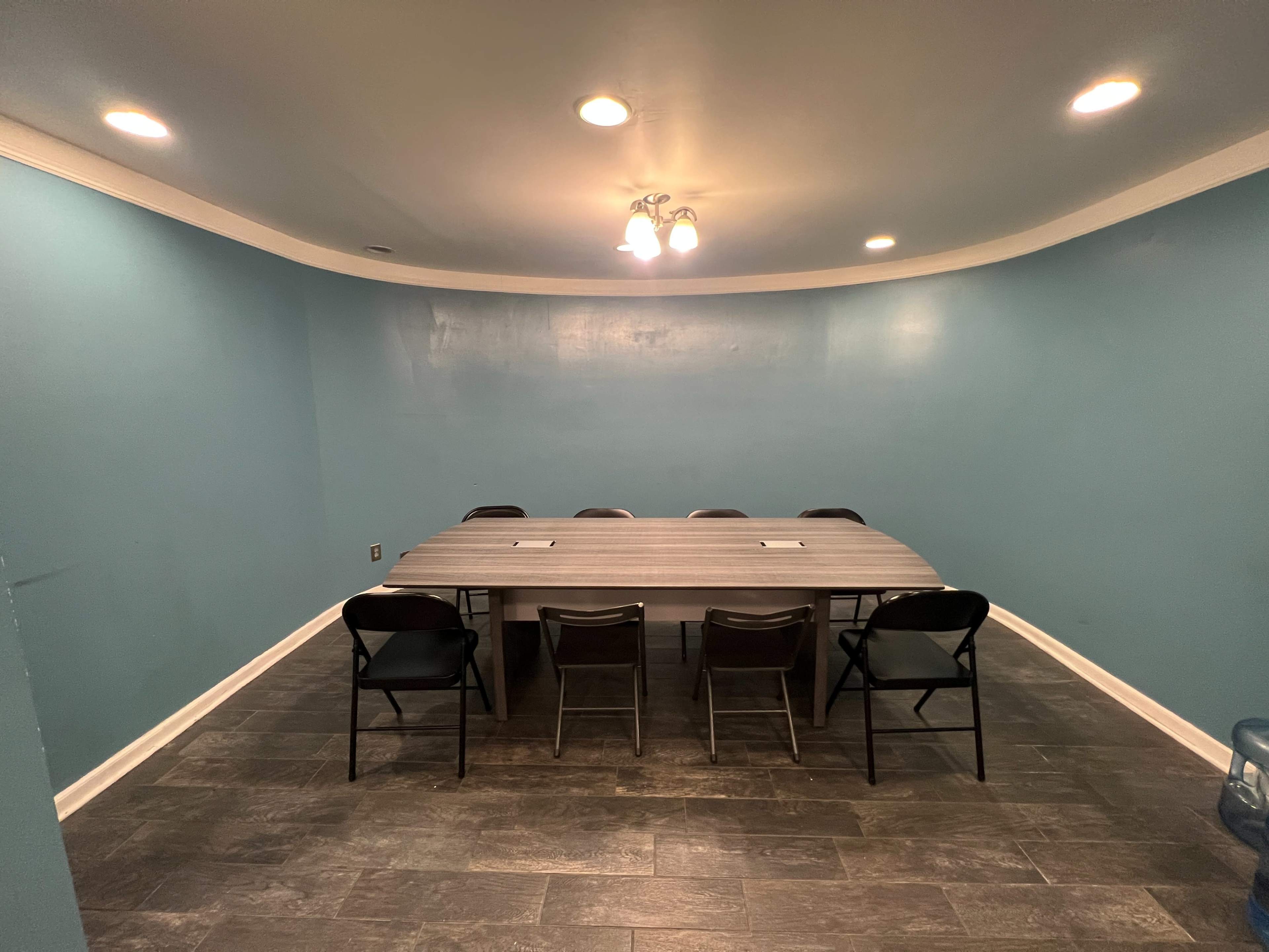A spacious meeting room features a large table surrounded by eight chairs, with blue walls and overhead lighting.