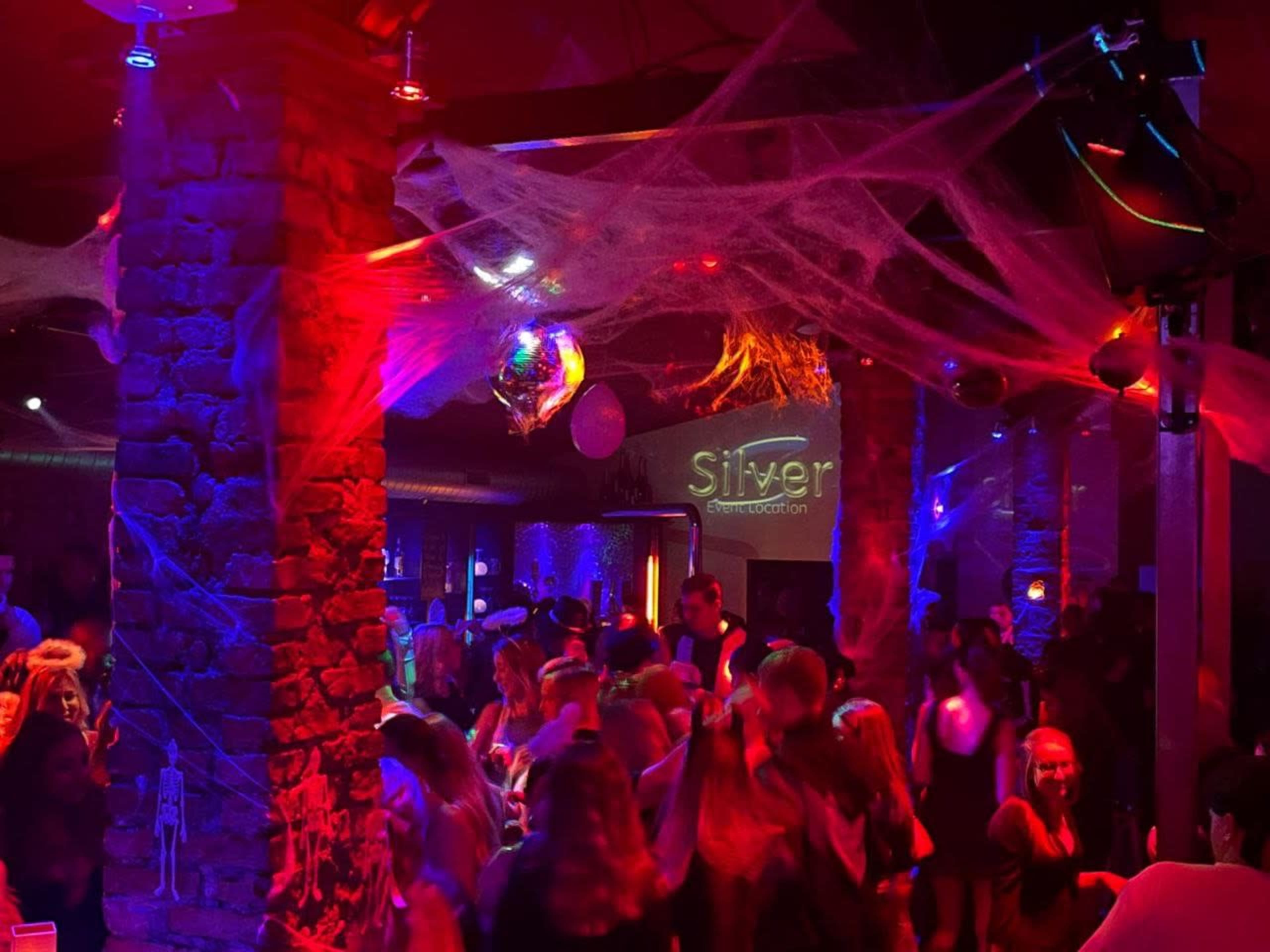 A crowded nightclub features colorful lights, disco balls, and Halloween decorations hanging from the ceiling.