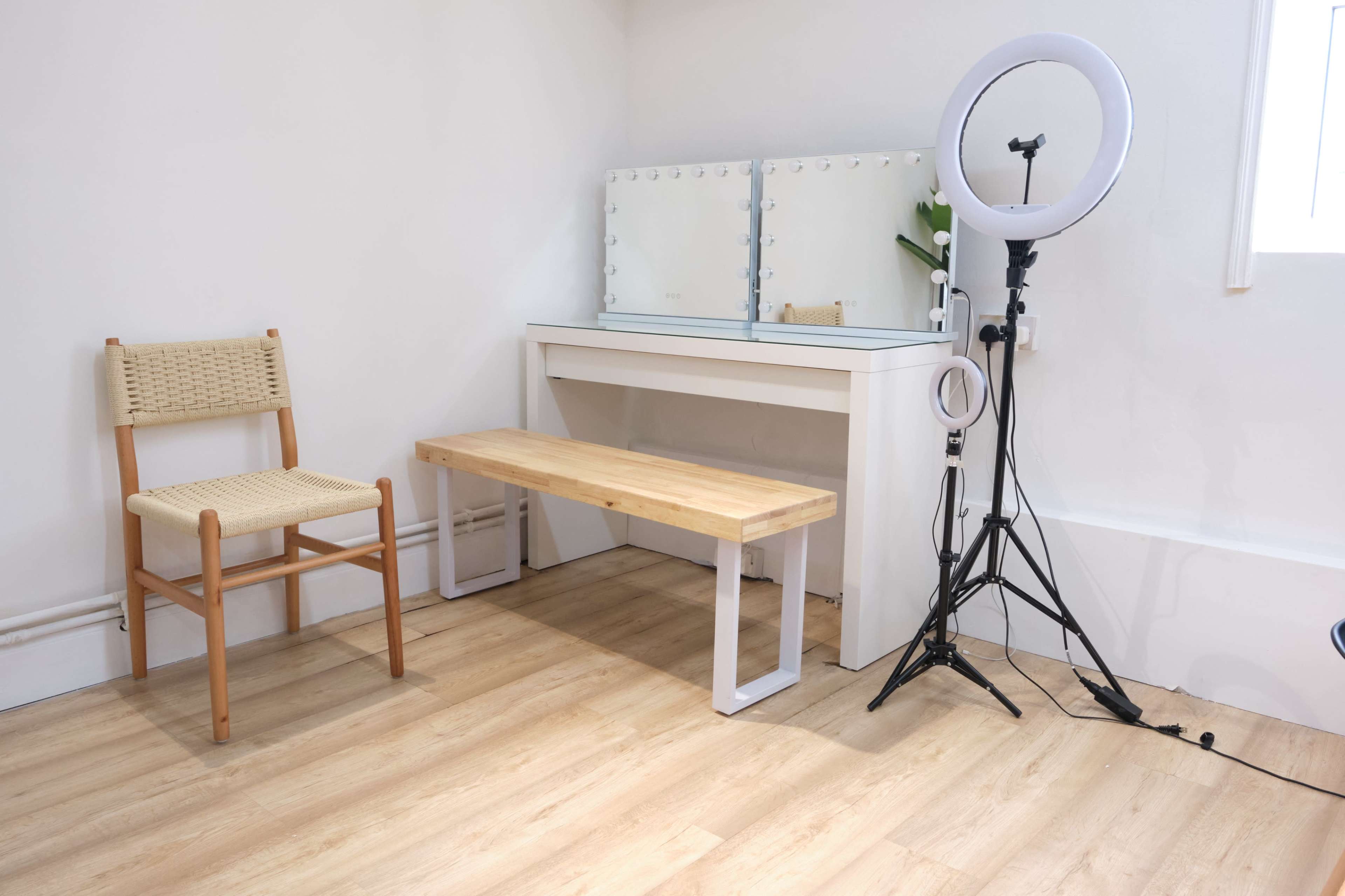 A simple vanity area with a mirror, a wooden bench, a chair, and a ring light on a stand in a brightly lit room with wooden flooring.