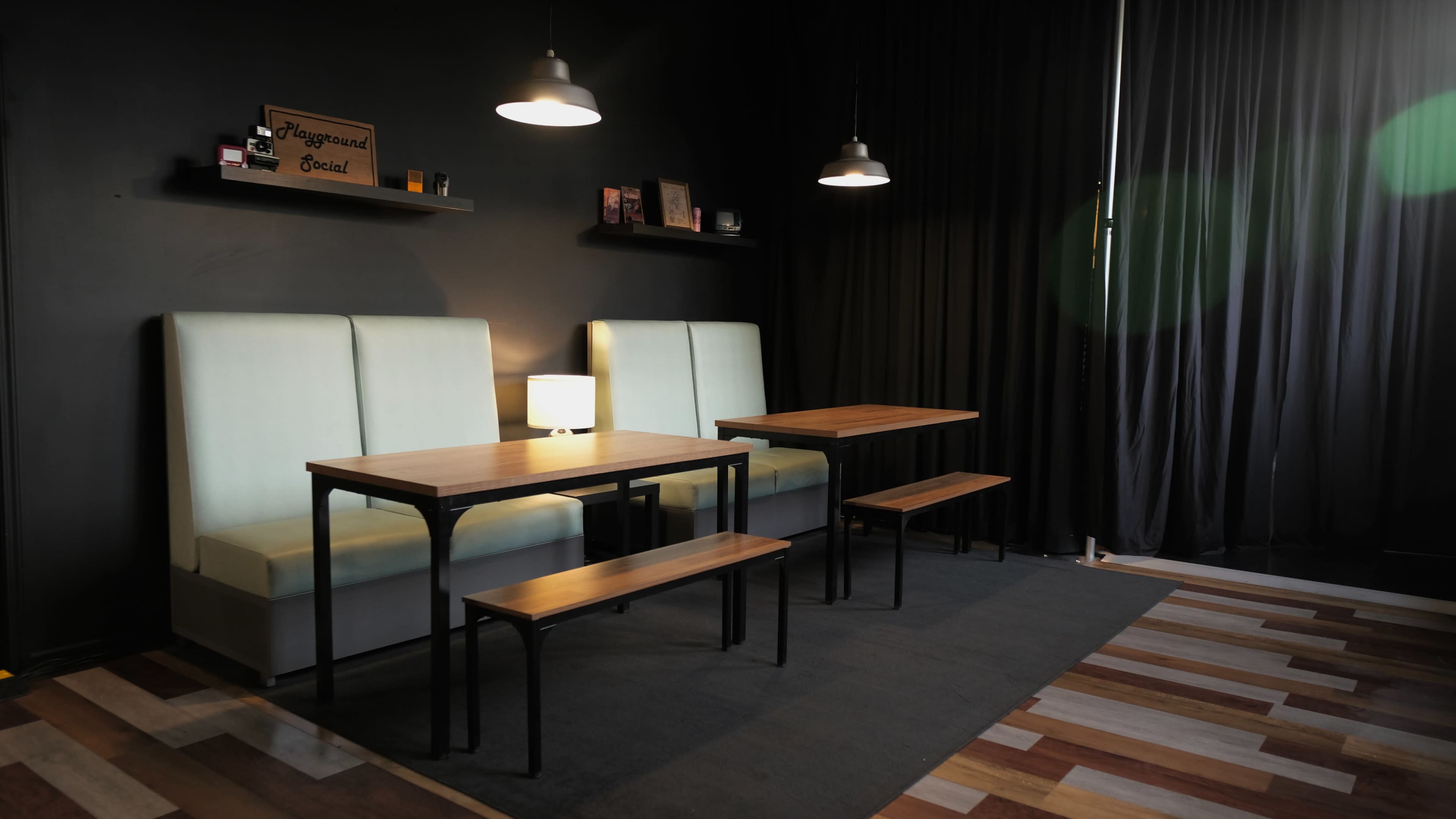 The image shows a minimalist café interior featuring a pair of light green upholstered booths, a wooden table, and two benches, with dark walls and soft lighting.