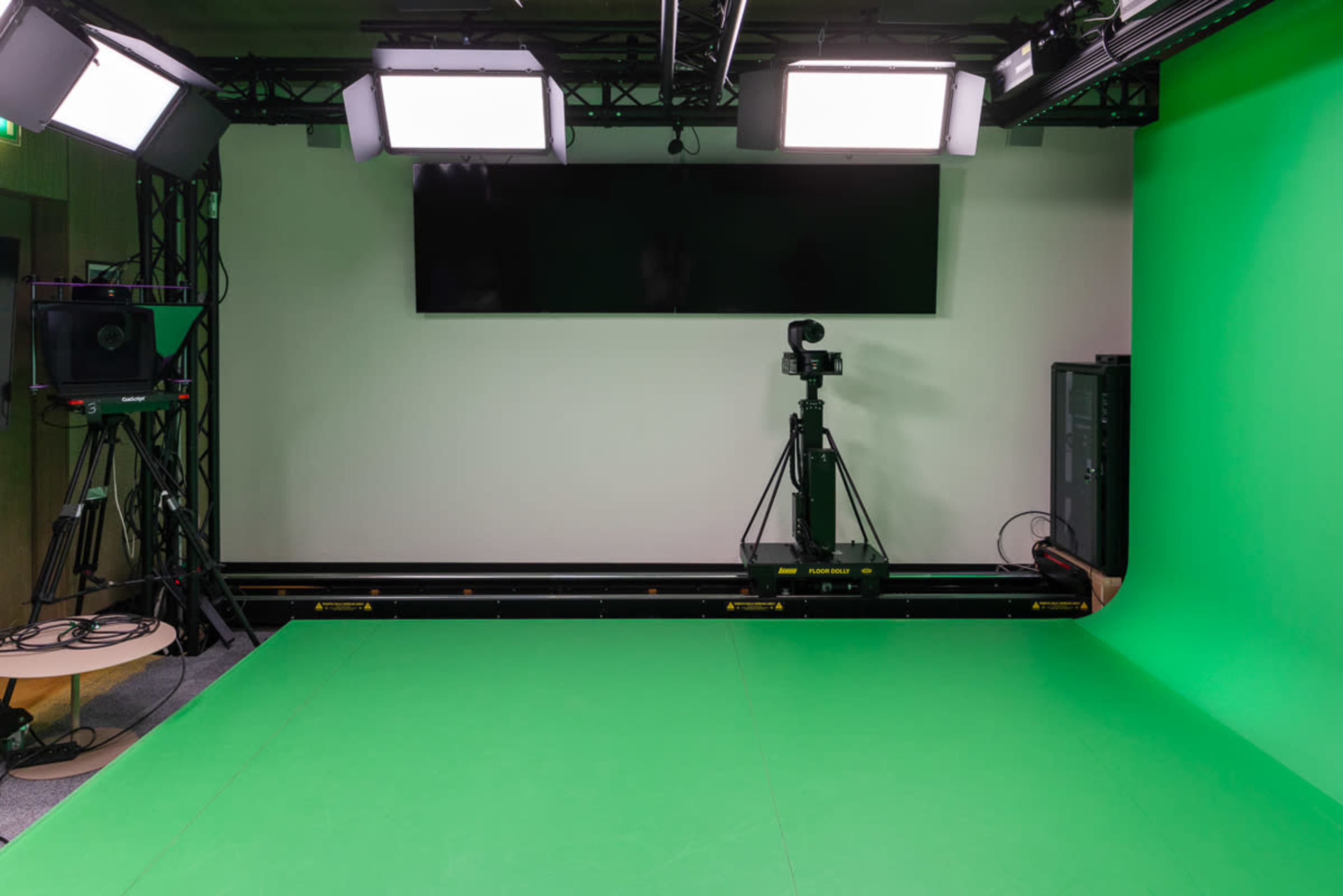 The image shows a green screen studio equipped with four overhead lights and a camera setup on a tripod.