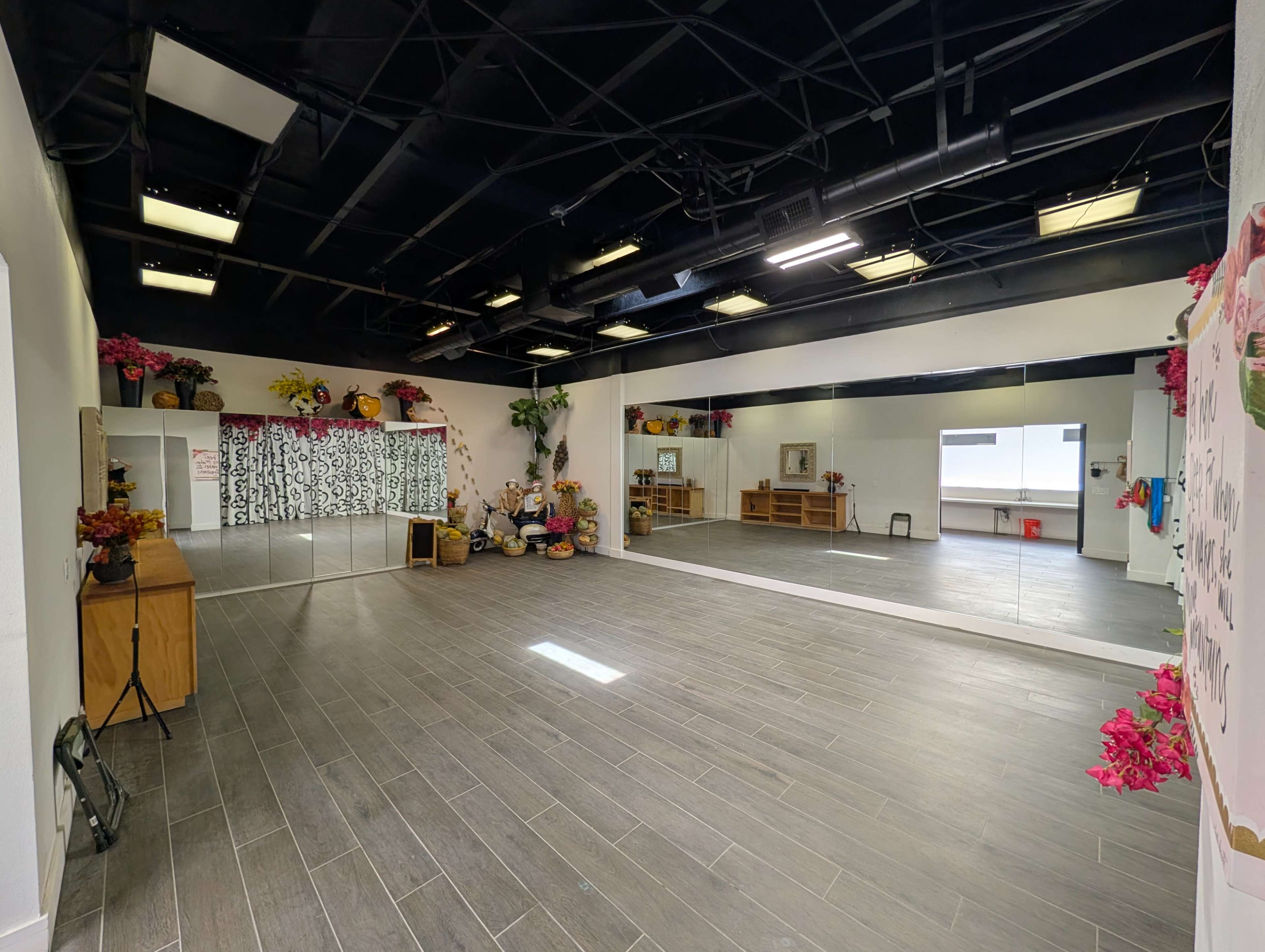The image shows a spacious studio with dark ceilings, gray floor tiles, and a large mirror along one wall, decorated with flowers and furniture.