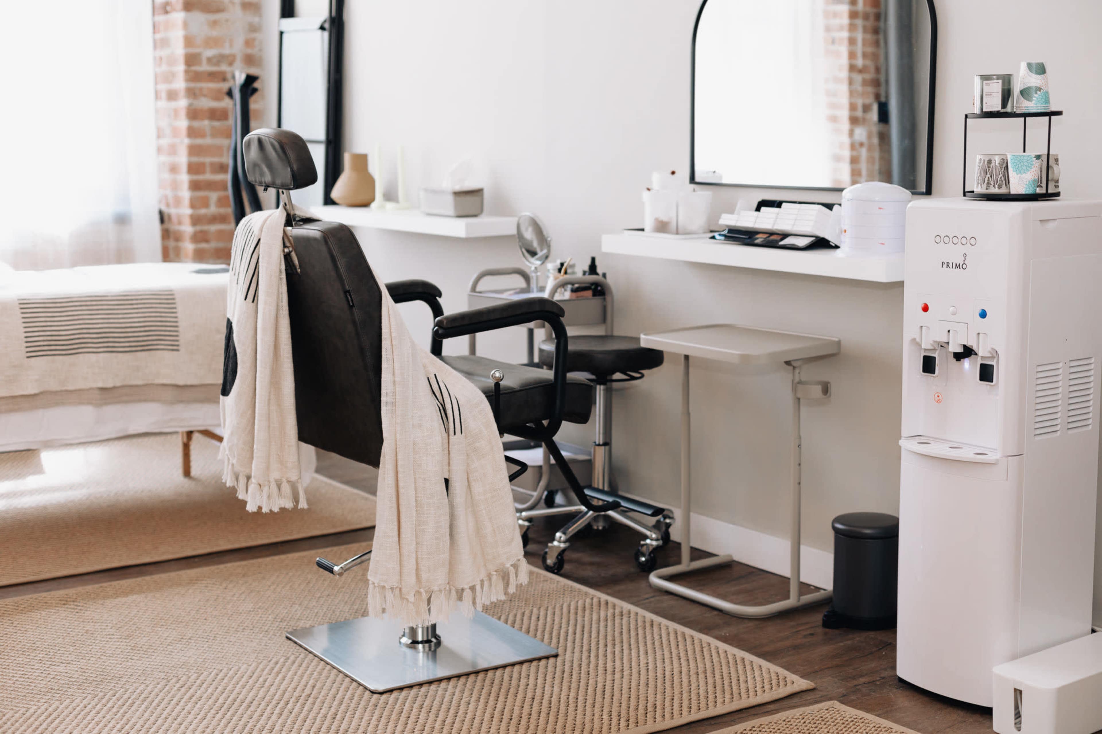 The image shows a minimalist salon interior with a barber chair, a mirror, a small table, and a water dispenser.