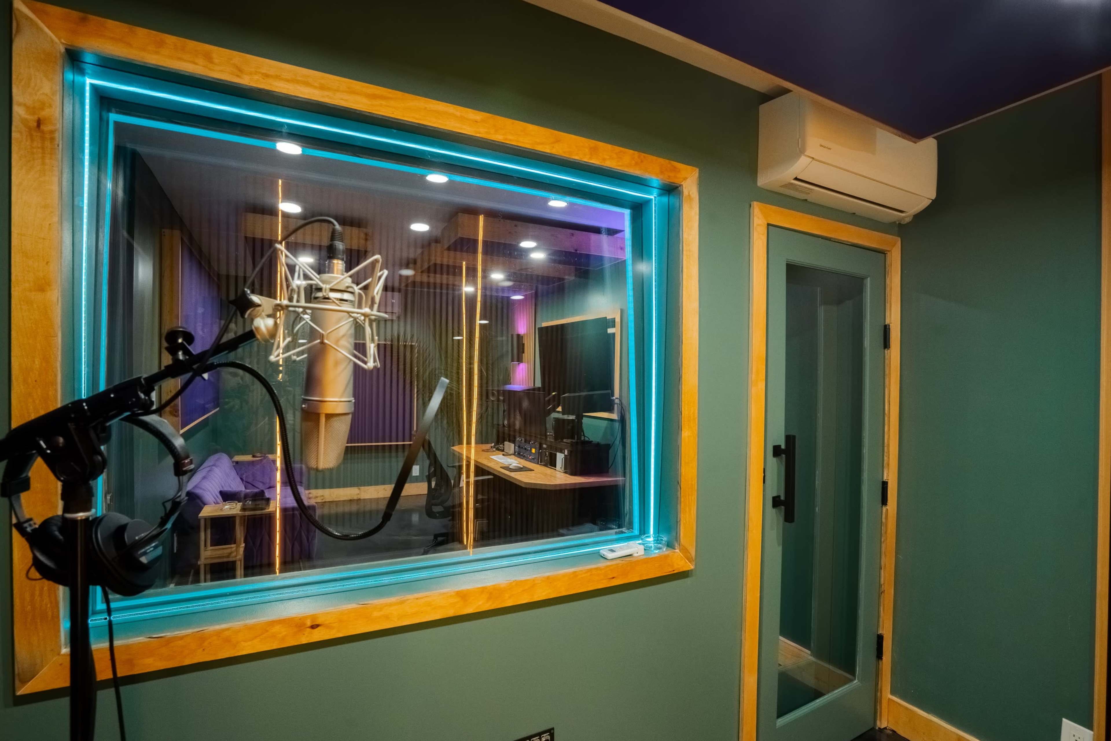 The image shows a soundproof recording studio with a microphone set up and a view into another room featuring sound equipment and a computer.