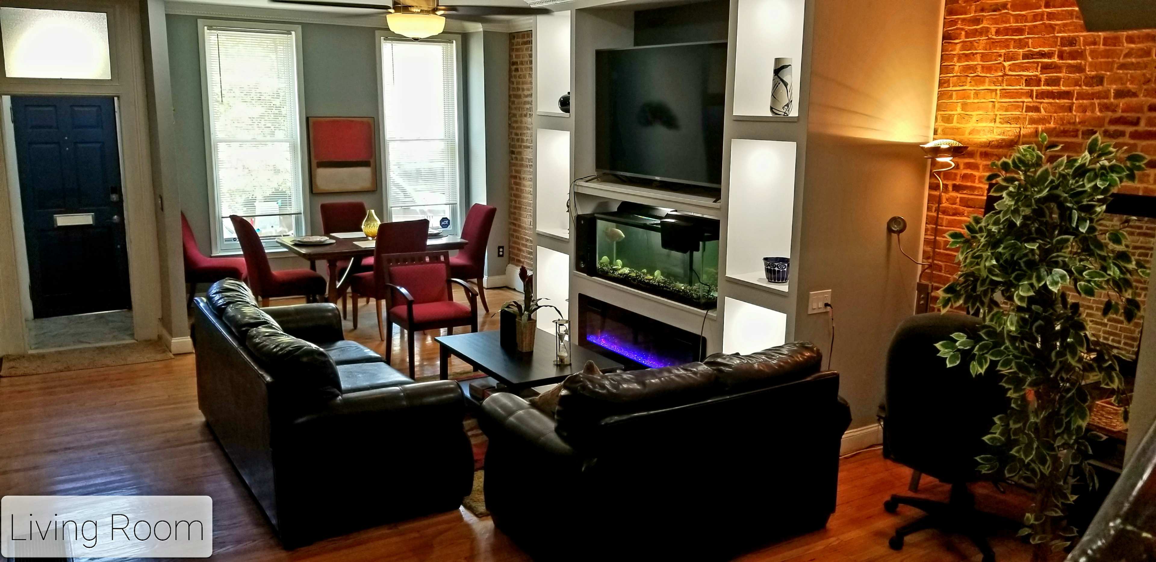 A modern living room features a dark sofa set, a dining area with red chairs, a television above a fireplace, and a large fish tank.