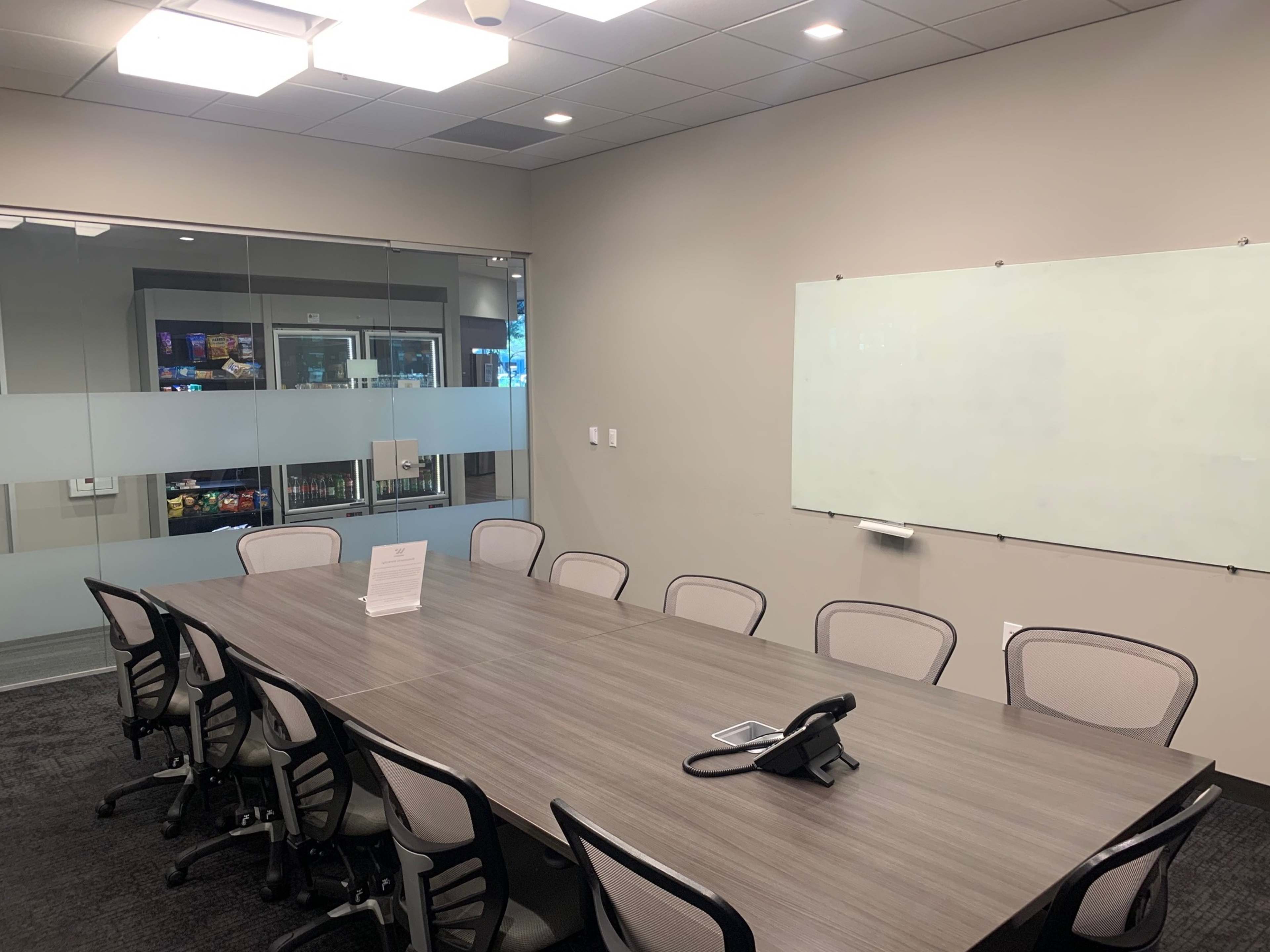 A conference room features a long wooden table surrounded by chairs, with a whiteboard on one wall and a glass partition showcasing a beverage fridge.
