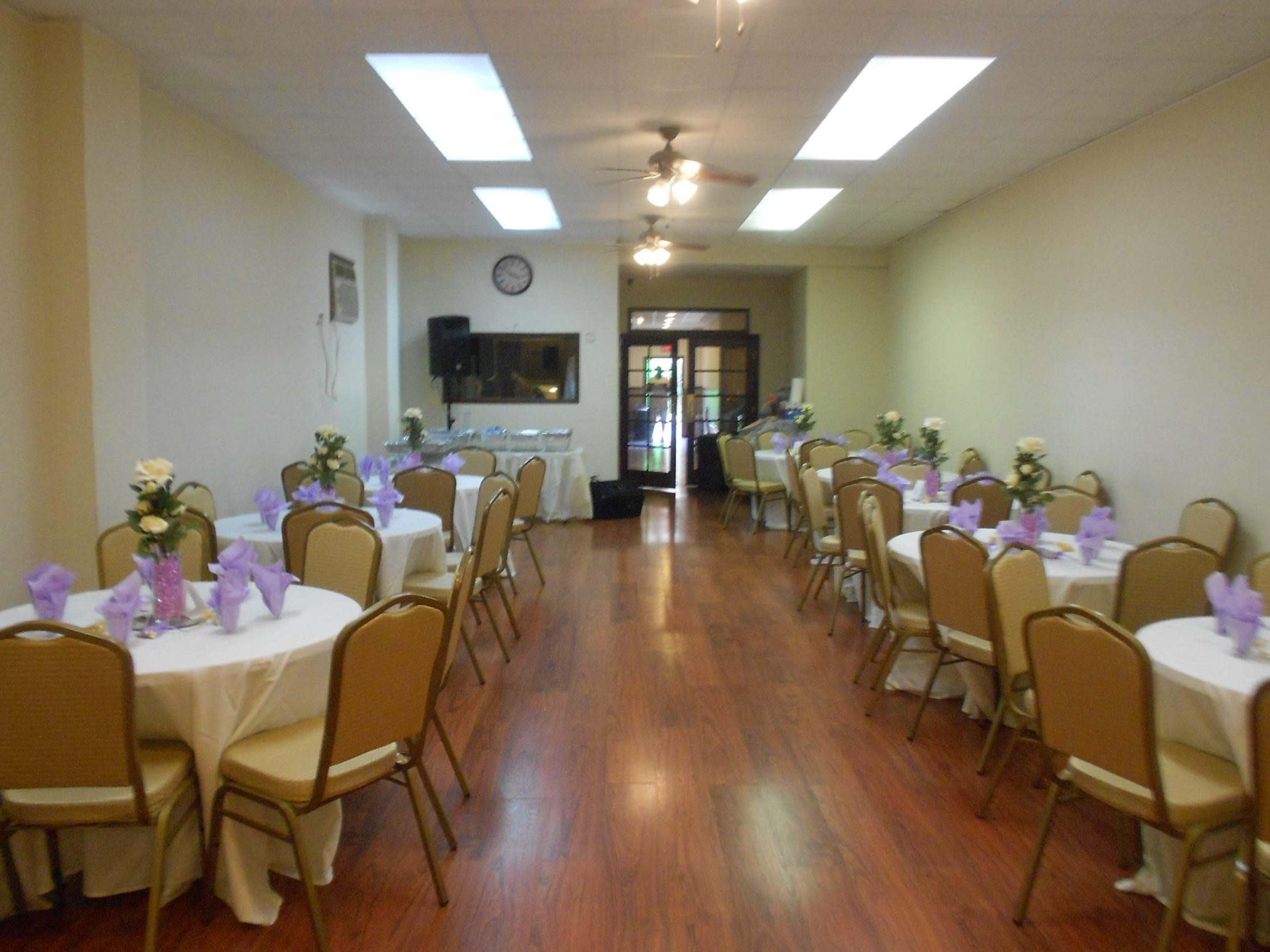 A banquet hall with round tables covered in white tablecloths and set with lavender napkins, arranged along a wooden floor.