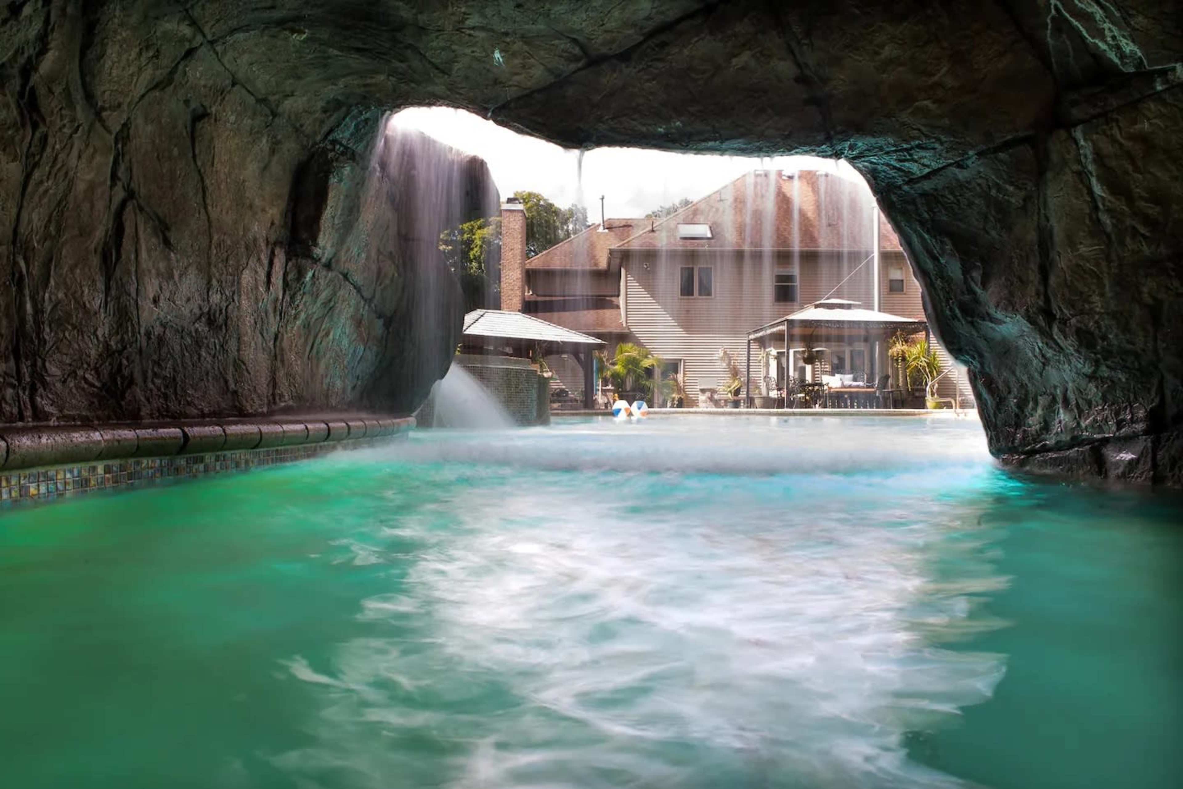 A large swimming pool with a cave-like rock formation, where water cascades down from the rocks into the pool, and a house is visible in the background.