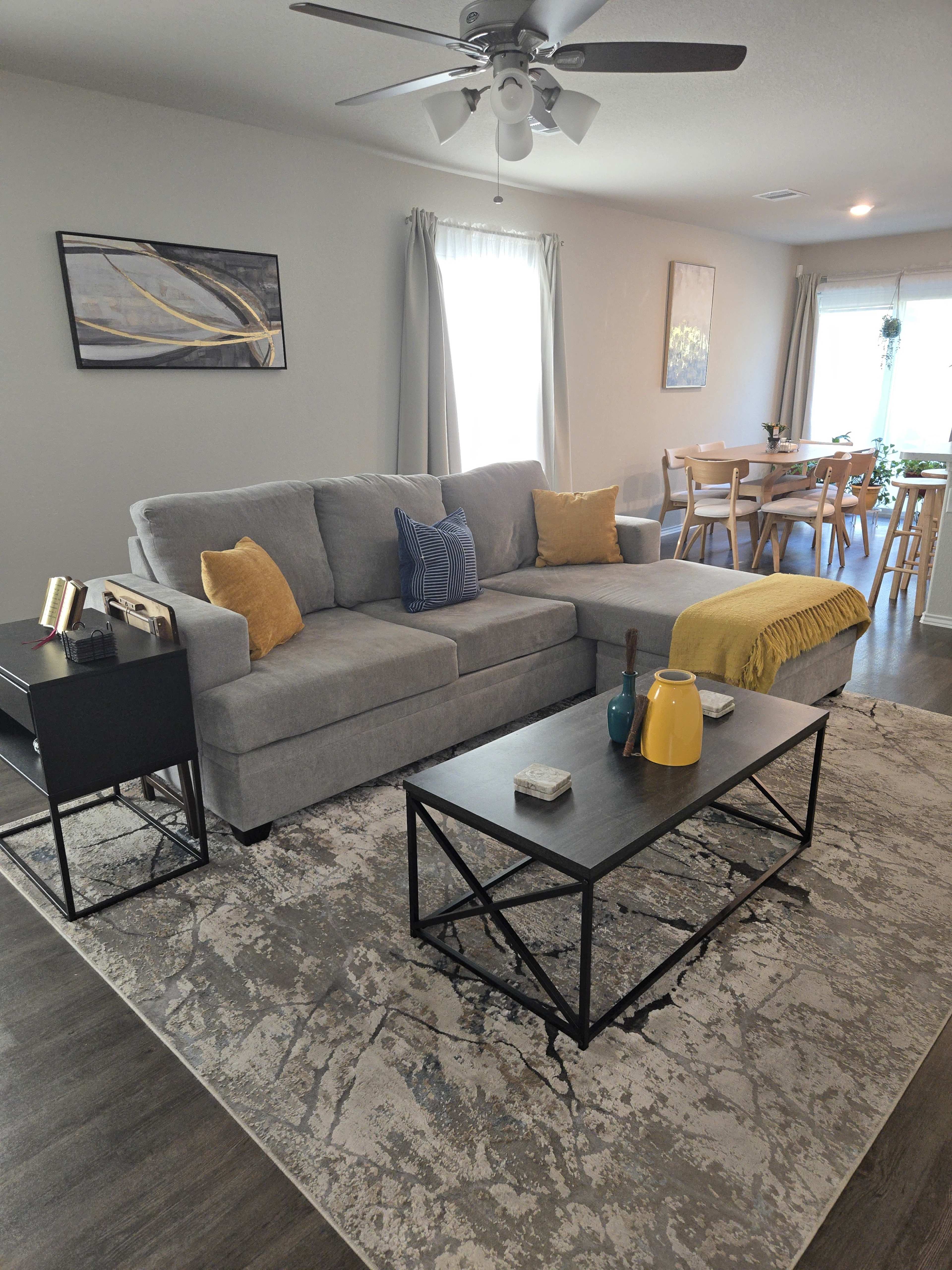 The image shows a living room with a gray sectional sofa, a black coffee table, decorative items, and a dining area in the background.