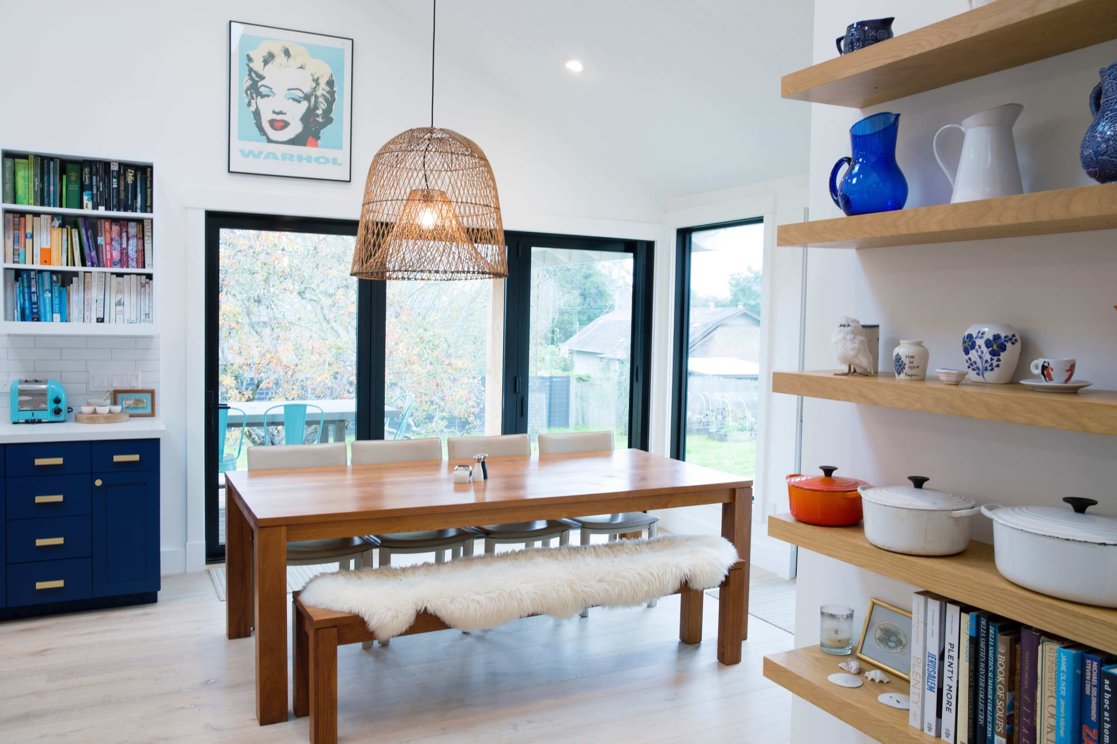 A modern dining area featuring a wooden table with a bench and chairs, pendant lighting, and shelves displaying various dishes and decorative items.