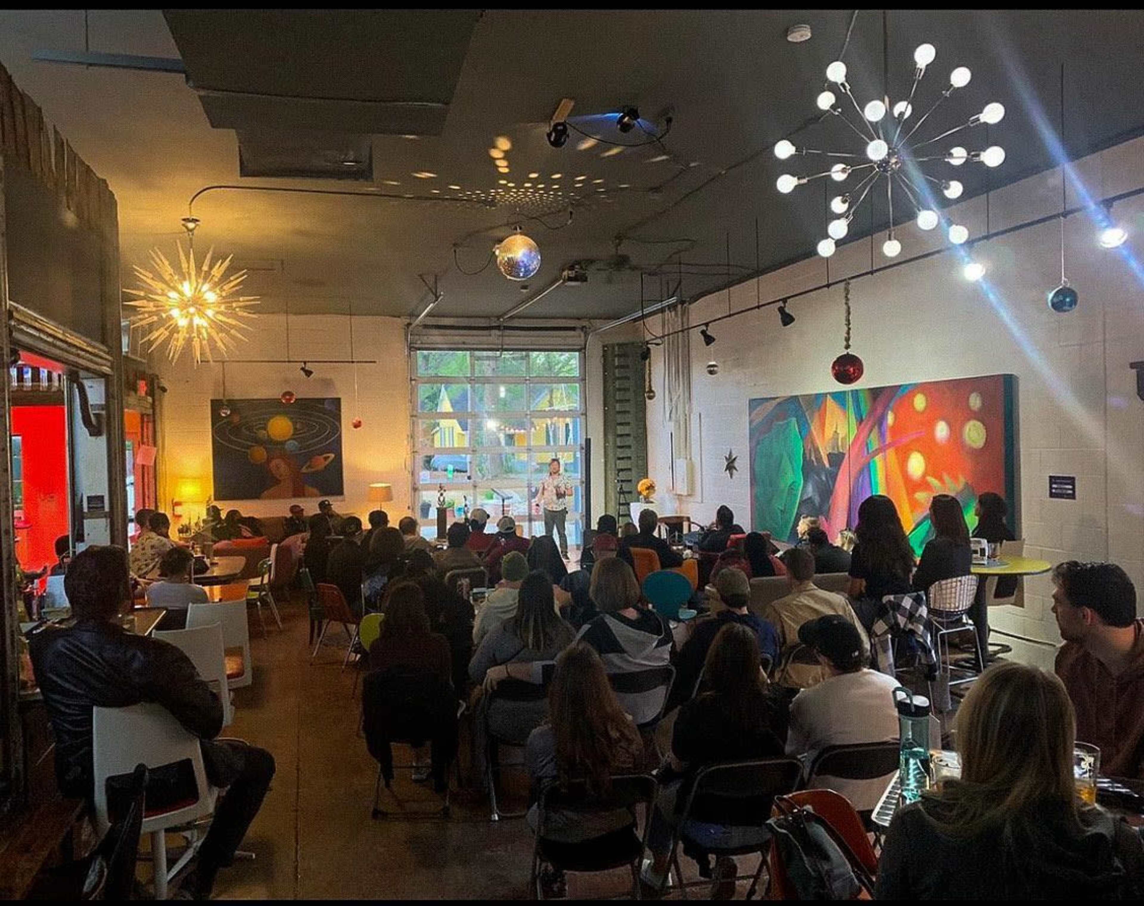 A crowd gathers in a lively indoor space with artistic decor, listening to a presentation on a stage.