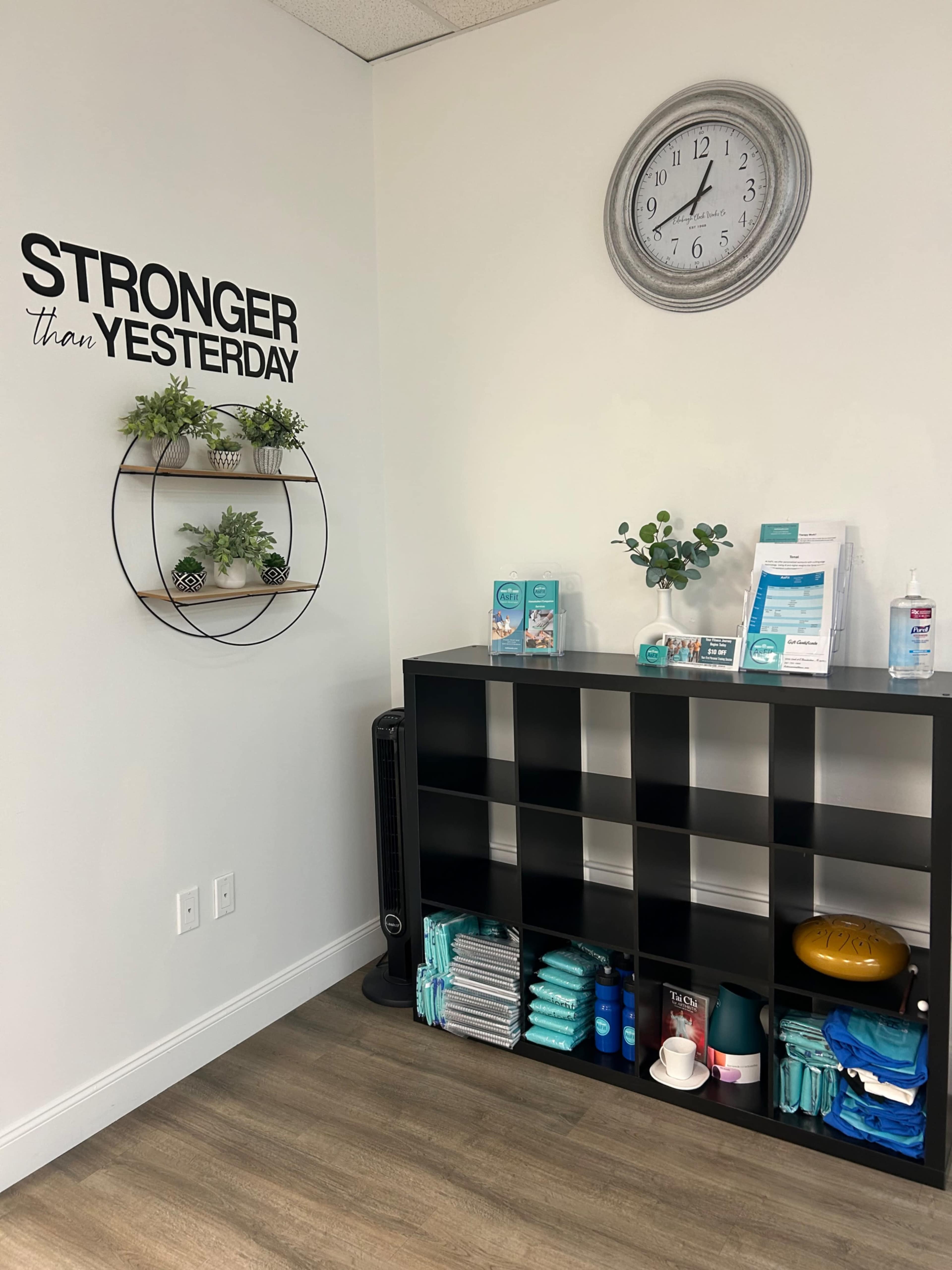 The image shows a reception area featuring a black shelving unit with organized items, a circular wall shelf with decorative plants, and a wall clock above a motivational quote.