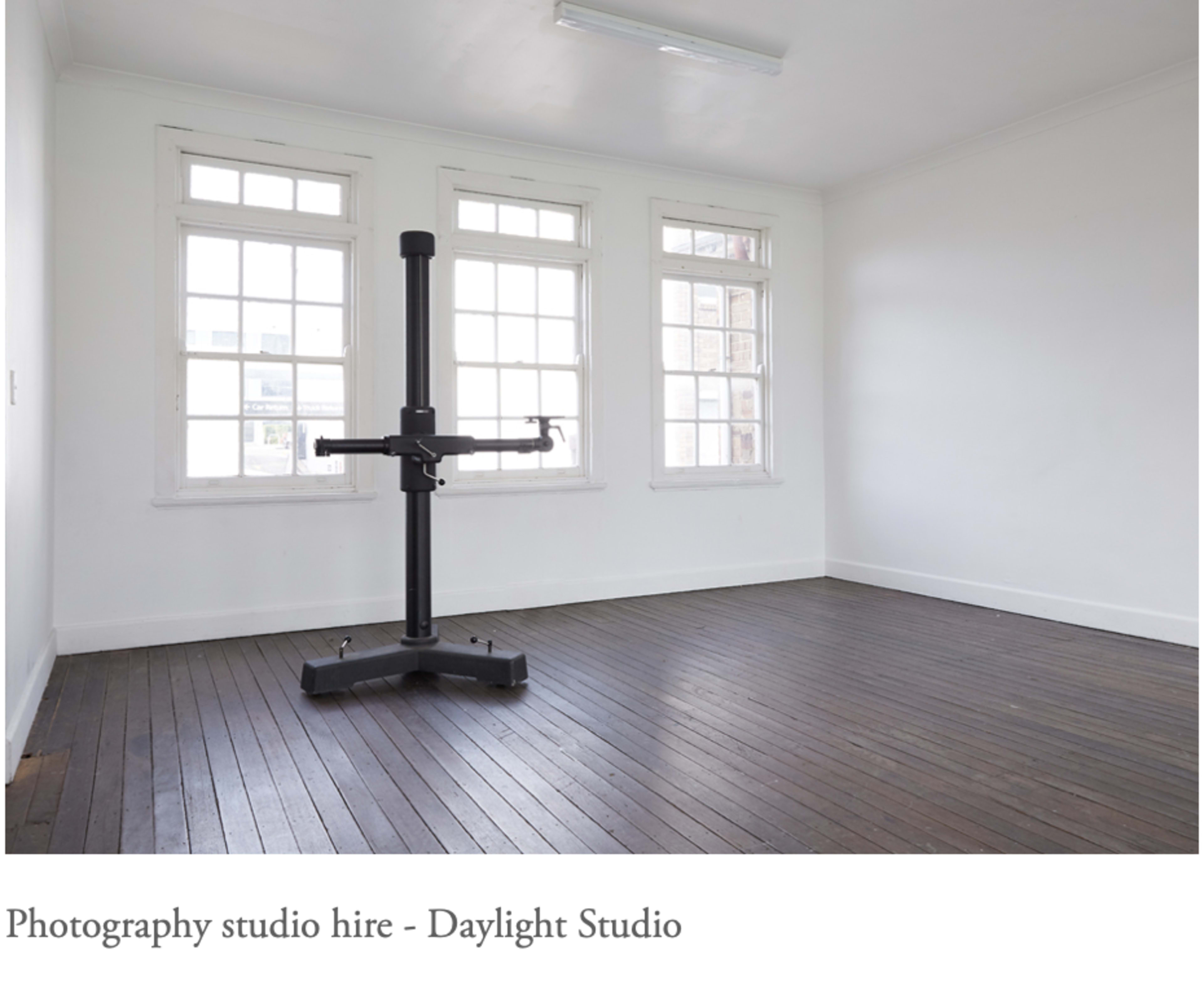 A minimally furnished photography studio with large windows and a central mounting stand on a wooden floor.