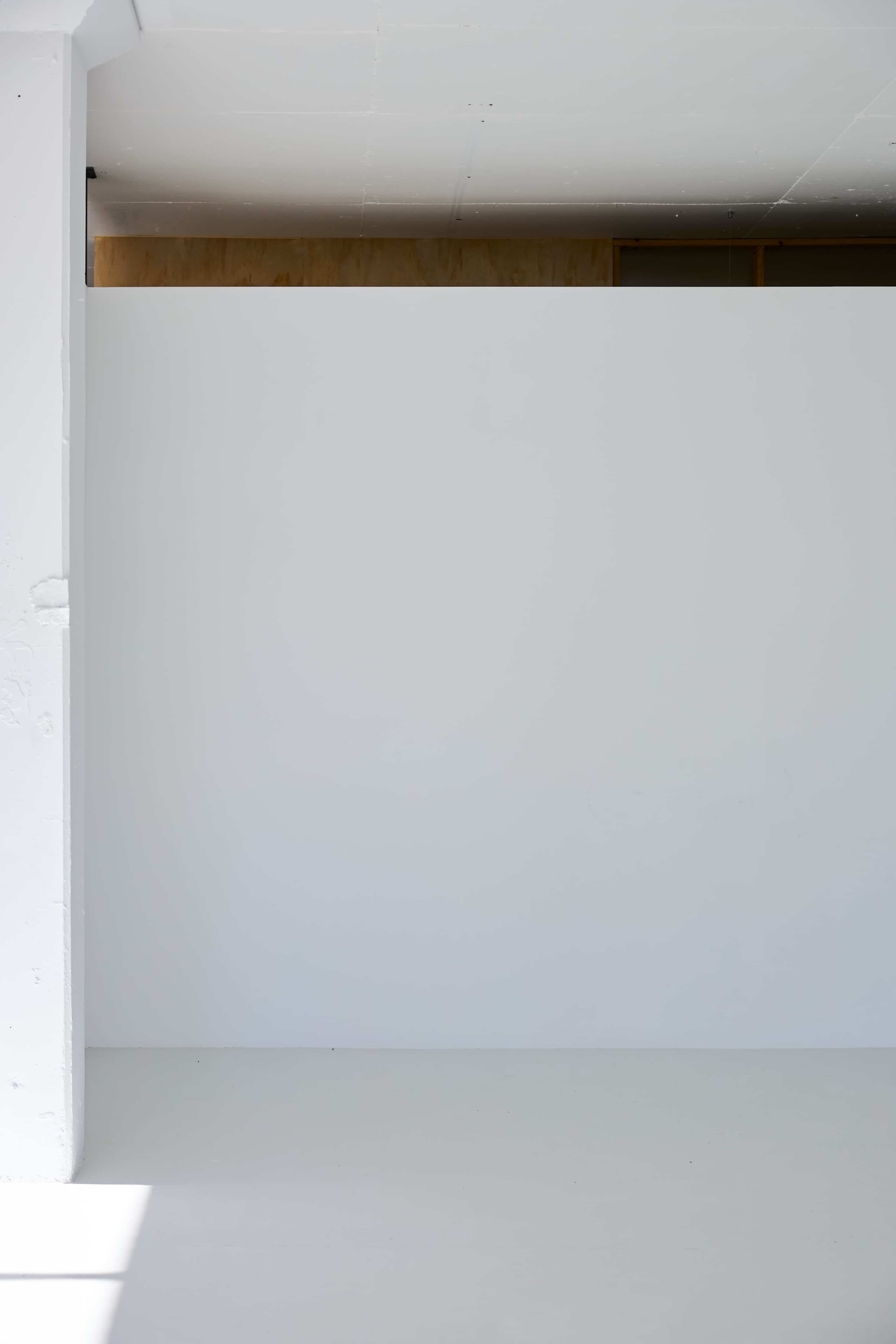 The image shows a minimalistic interior space featuring a plain white wall and a light-colored floor.