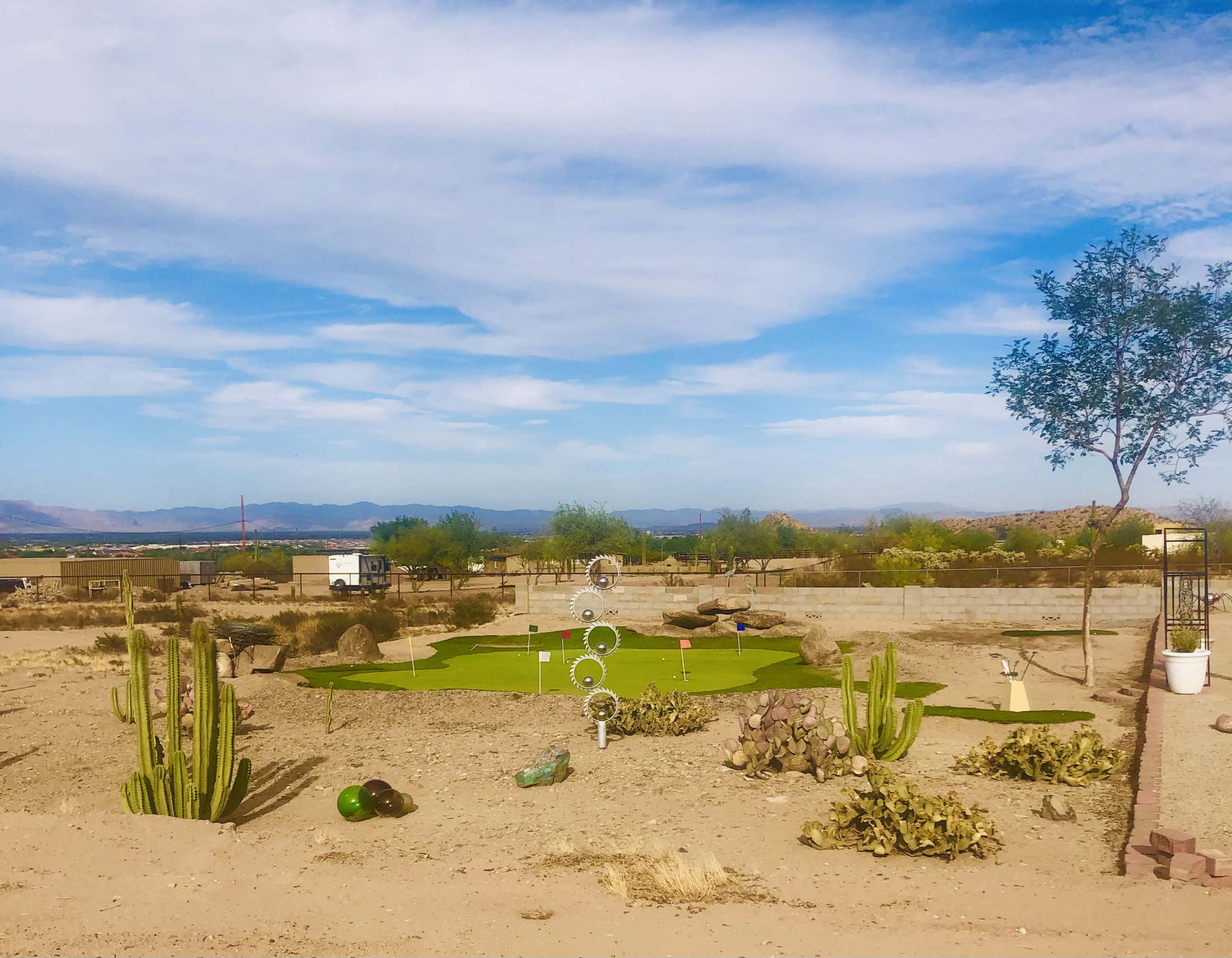 A small putting green surrounded by desert landscaping, cacti, and scattered boulders under a blue sky with clouds.