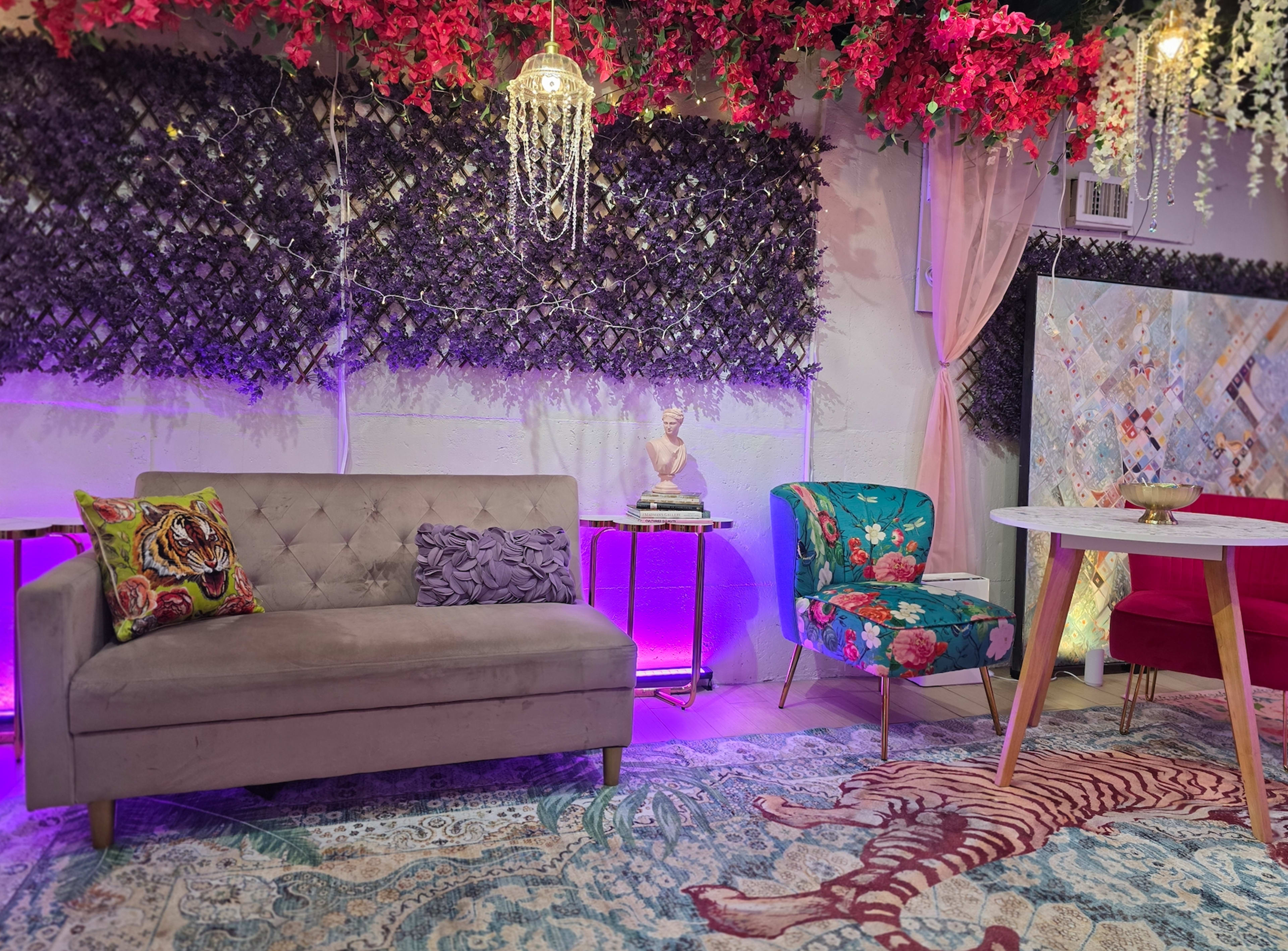 The image shows a vibrant interior space featuring a grey sofa, colorful chairs with floral patterns, a round table, and decorative elements including flowers, a chandelier, and a patterned rug.