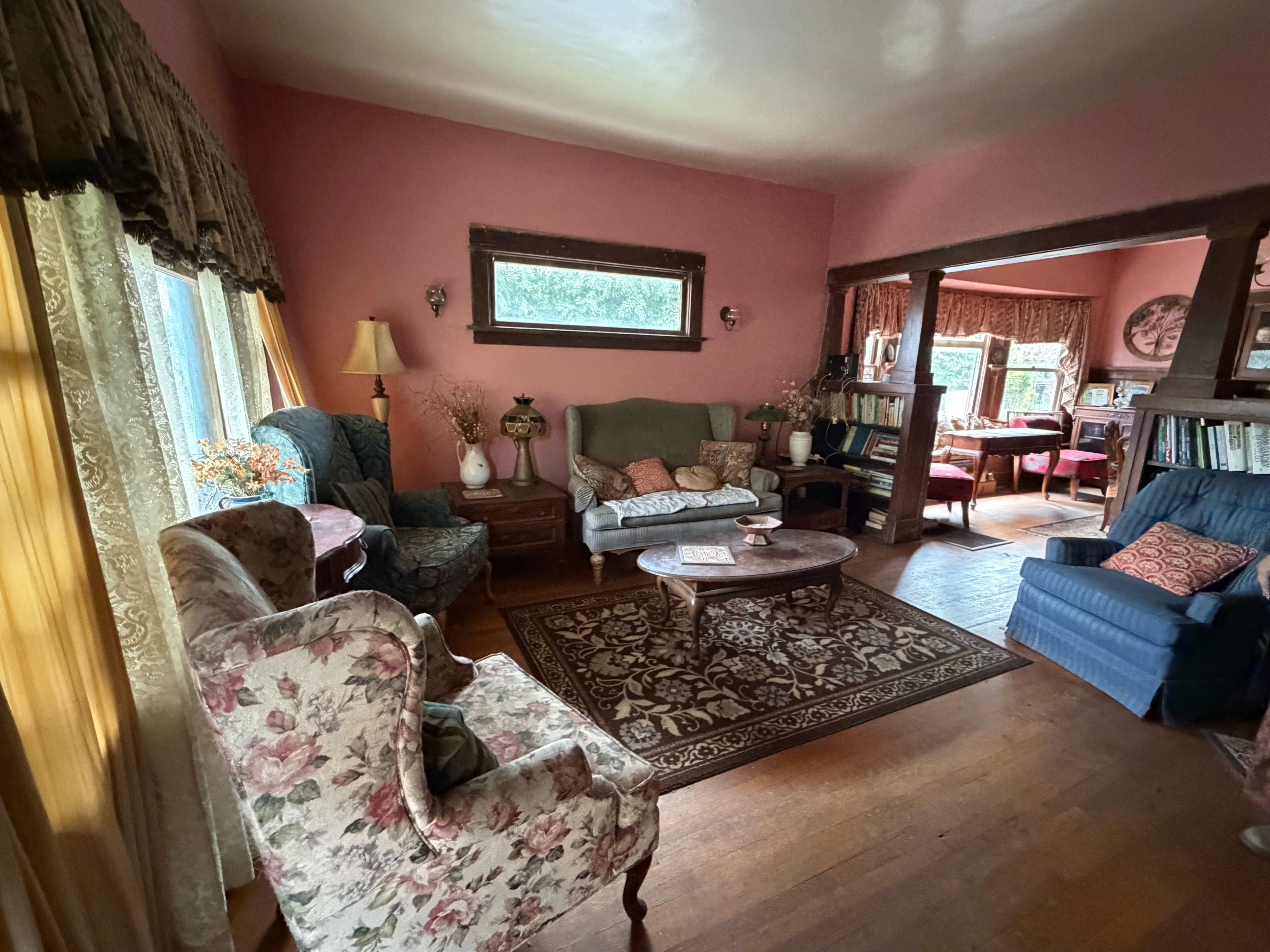 The image shows a cozy living room with floral-patterned chairs, a central coffee table, and a view into a dining area with additional furniture.