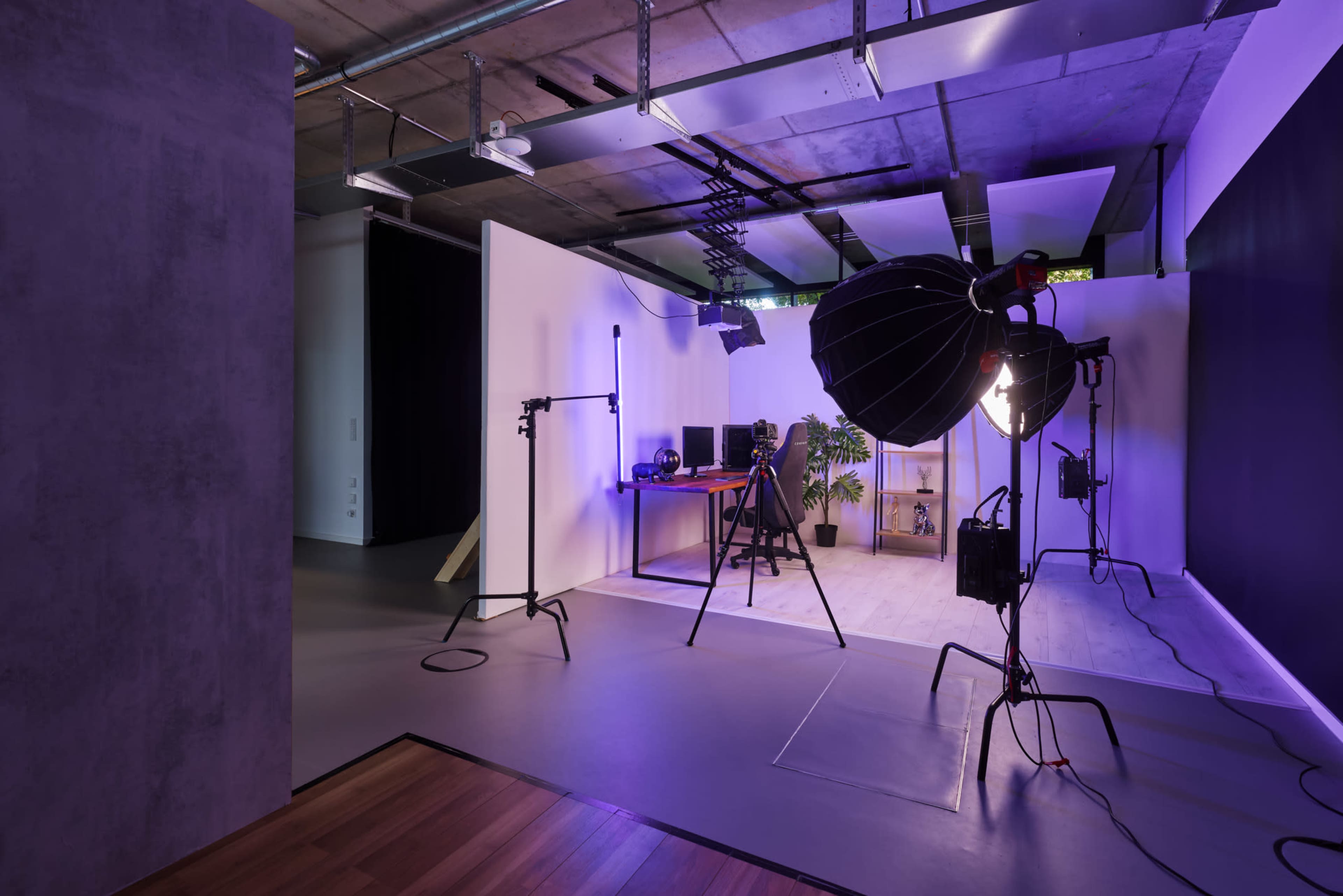 A modern photography studio featuring various lighting equipment, a work desk with computers, and a large potted plant.