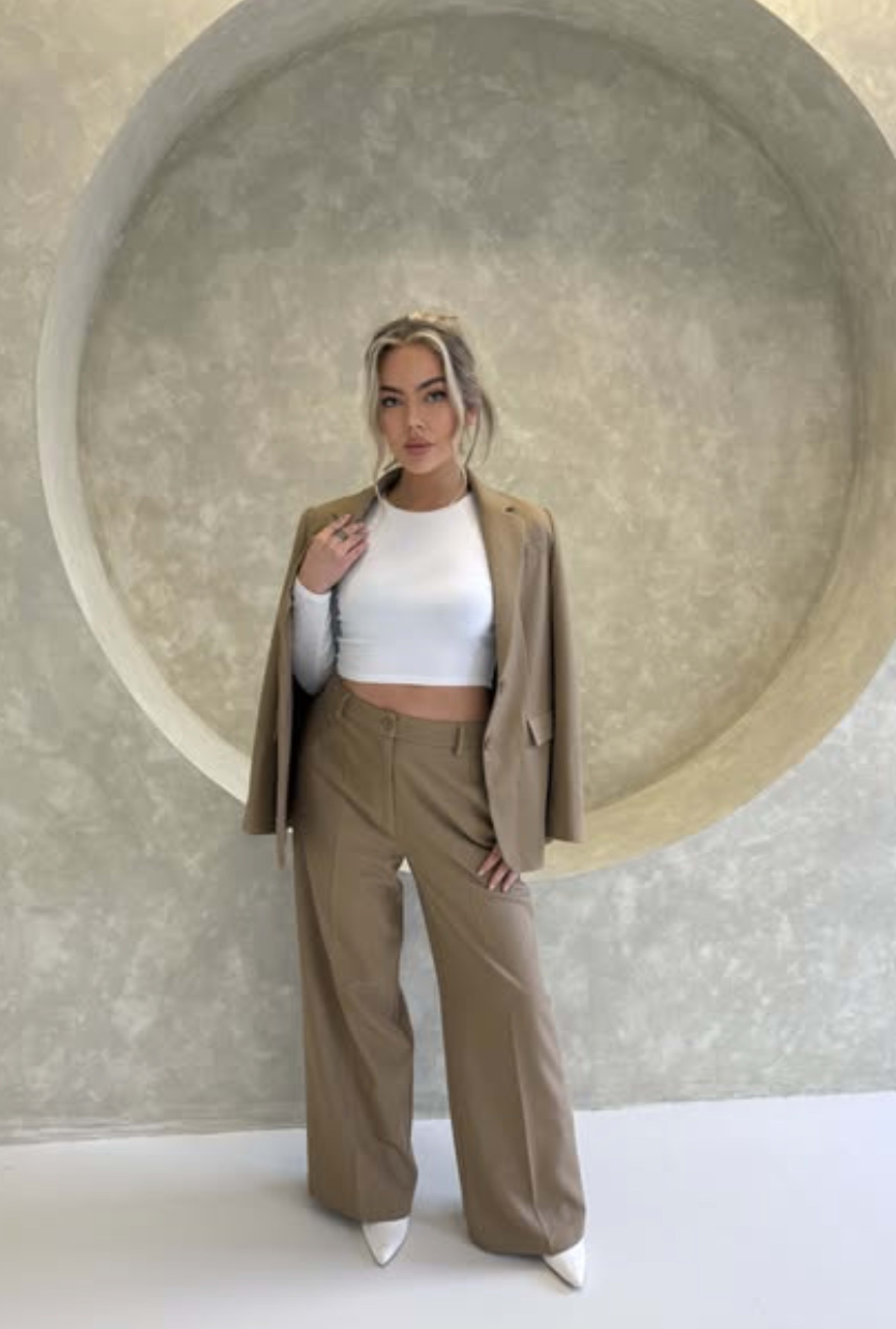 A woman stands in front of a textured circular backdrop, wearing a beige suit with a cropped white top and pointed-toe heels.