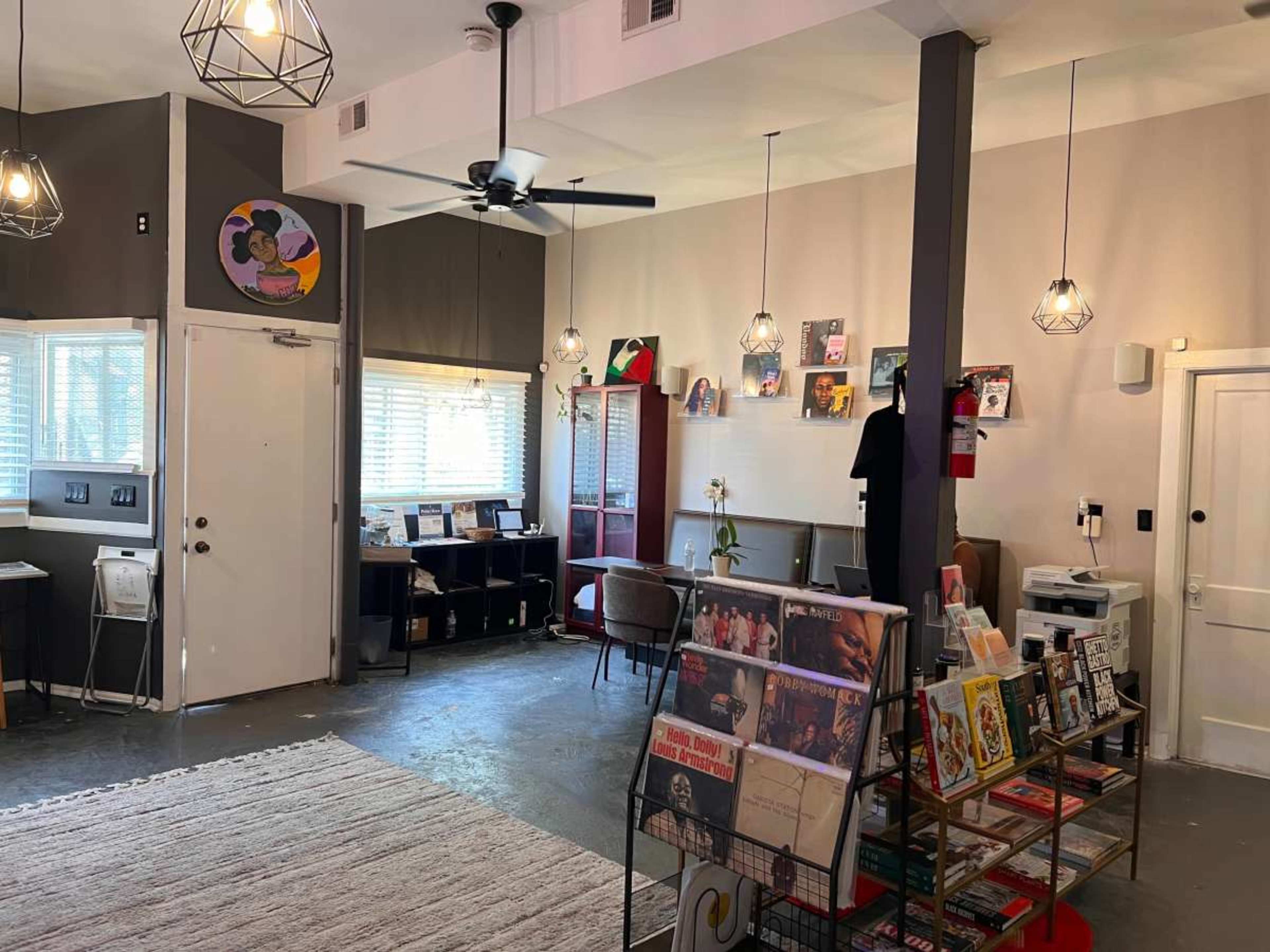 The image shows a modern, well-lit interior space featuring a reception area, various art displays, a sitting area, and a rack of magazines and books.