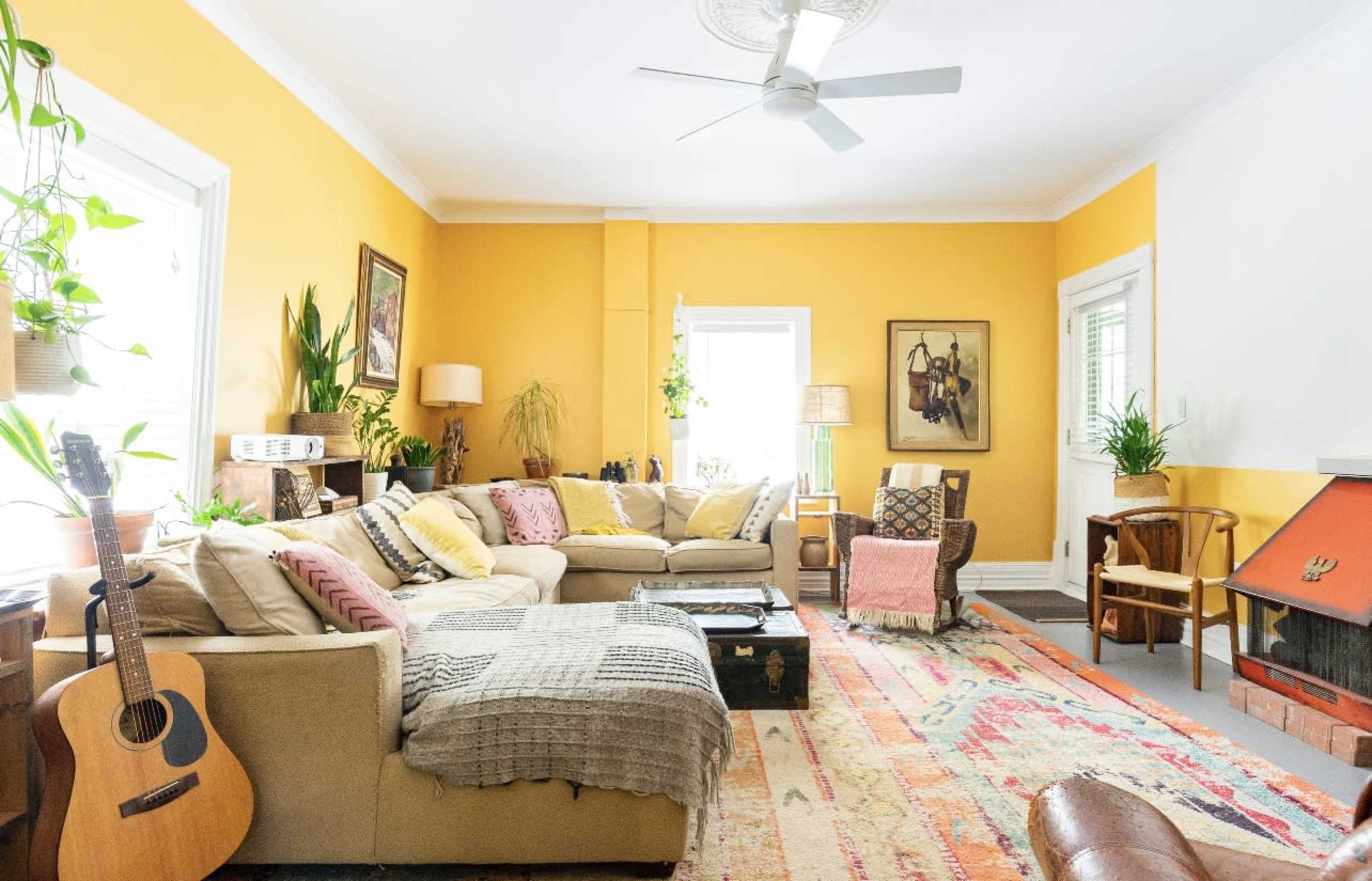 A cozy living room features a yellow wall, a large sectional sofa with various textured throws, an acoustic guitar, and a vintage rug.