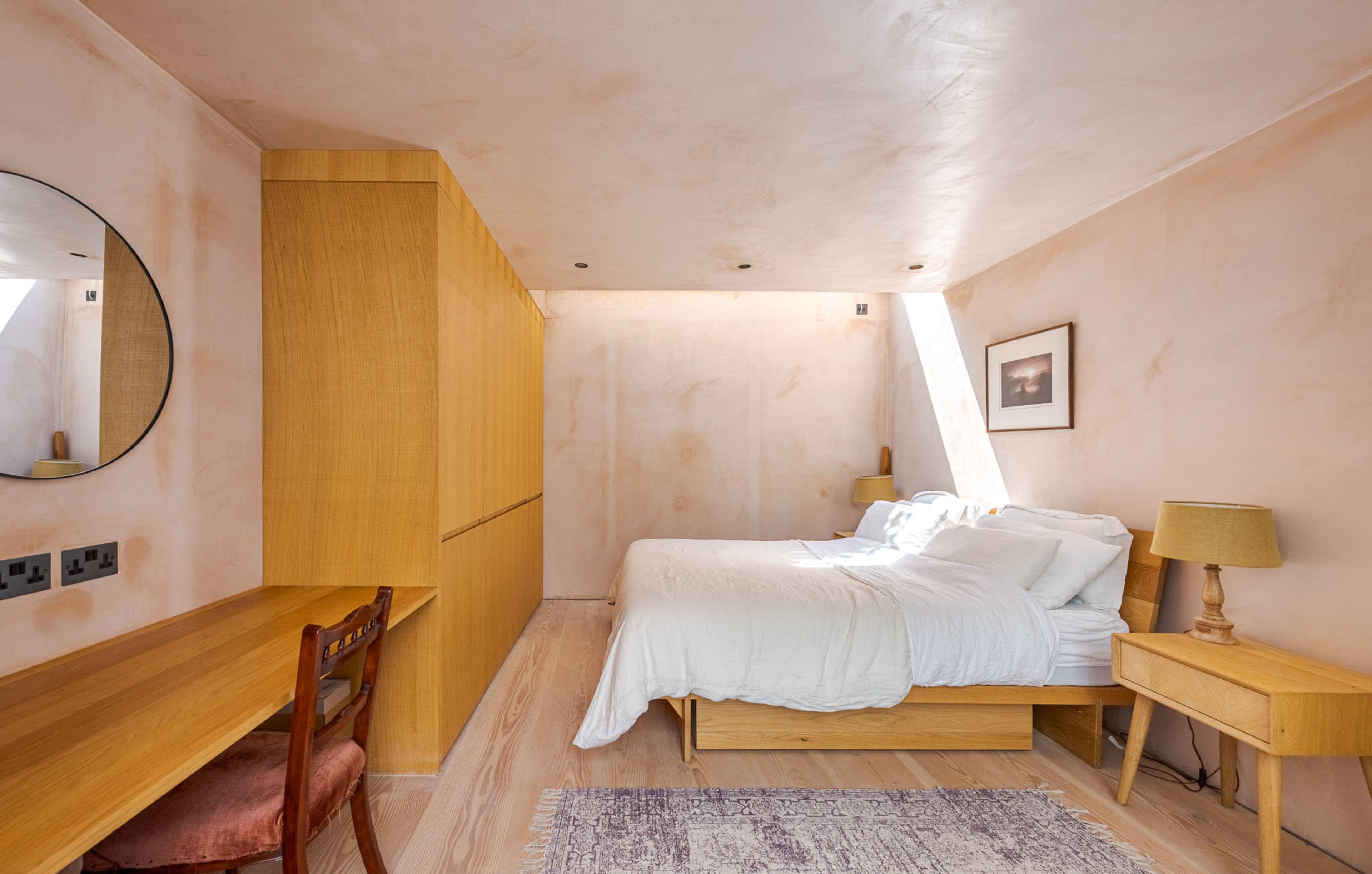 A minimalist bedroom features a bed, wooden furniture, and light-colored walls with natural lighting from a window.