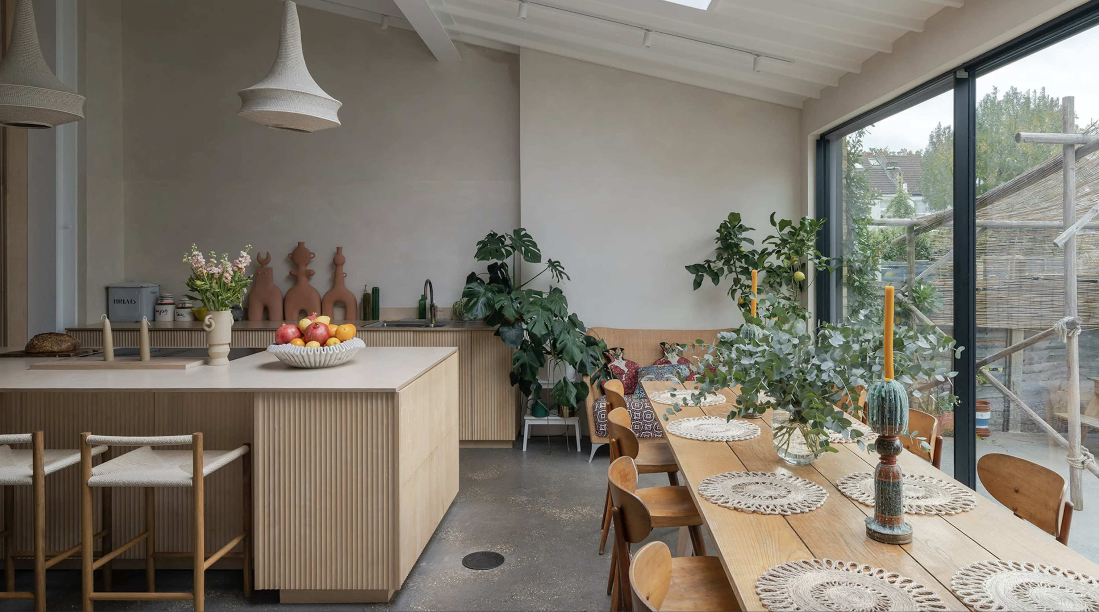 A modern kitchen features a wooden dining table, plants, and a counter with fruit, illuminated by natural light from large windows.