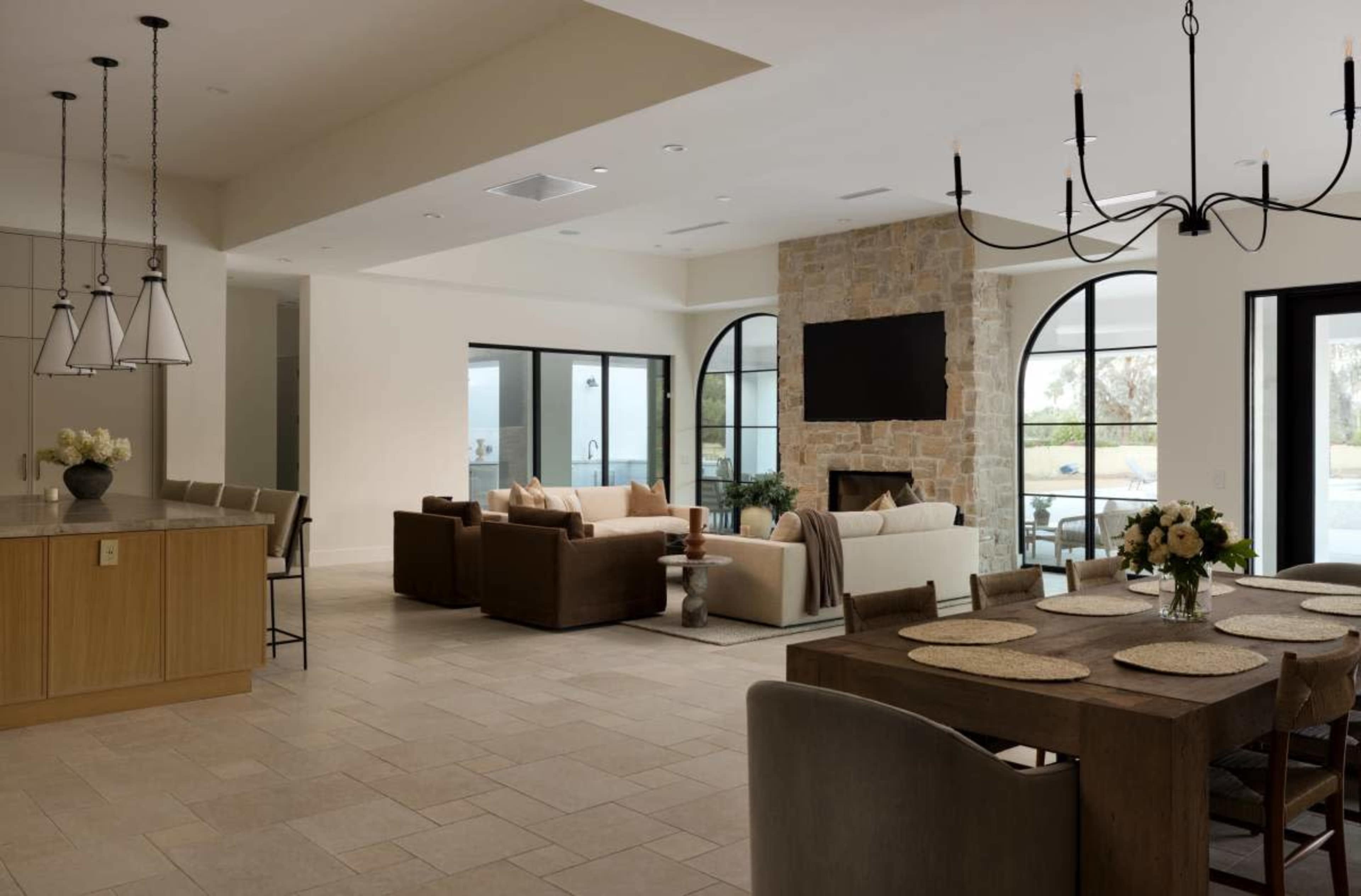 The image shows an open-concept living area featuring a dining table, a seating area by a stone fireplace, and large windows allowing natural light to enter.