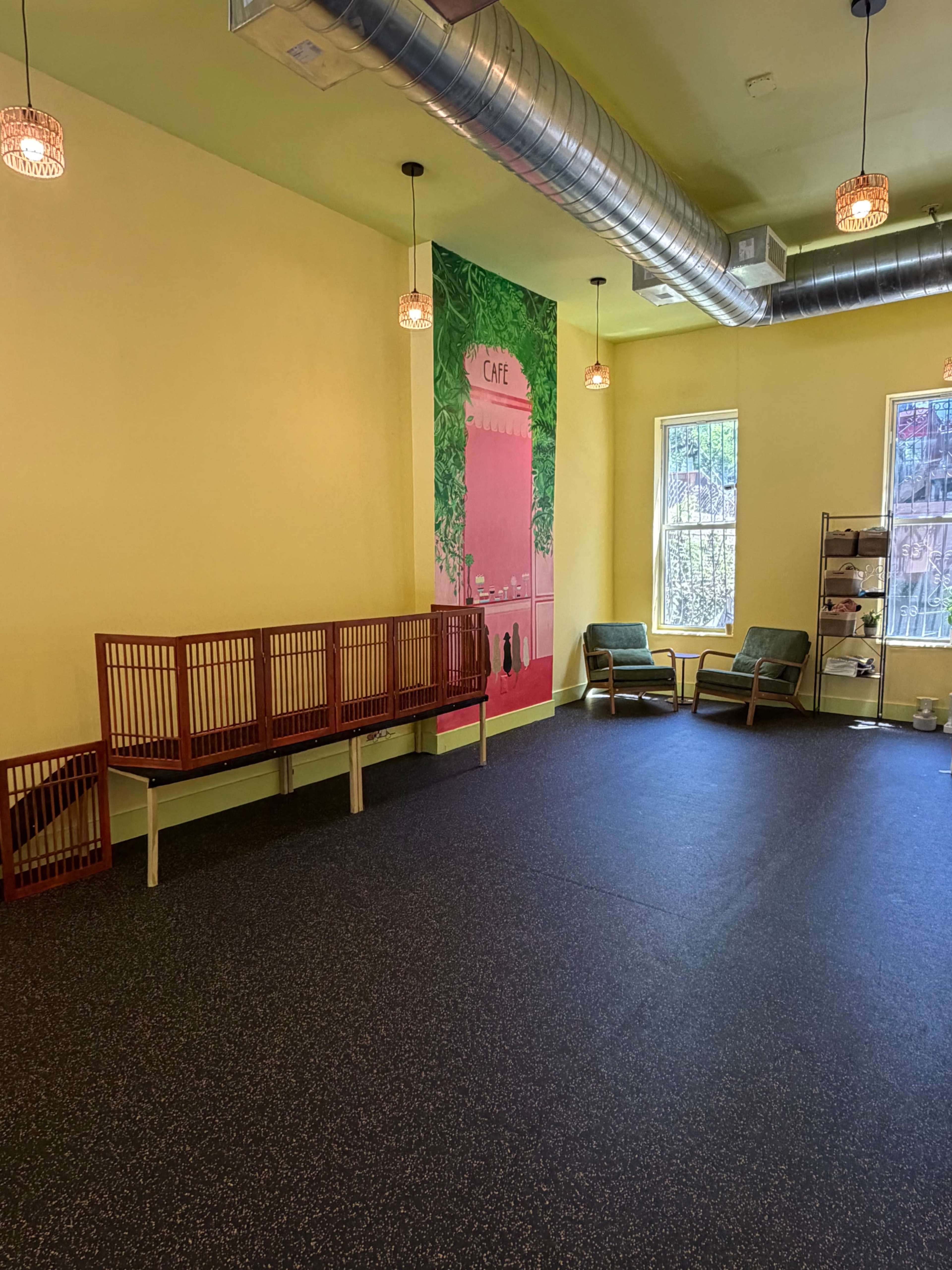 The image shows a spacious interior with yellow walls, a mural depicting a café, wooden seating, and two green chairs beside a shelf.