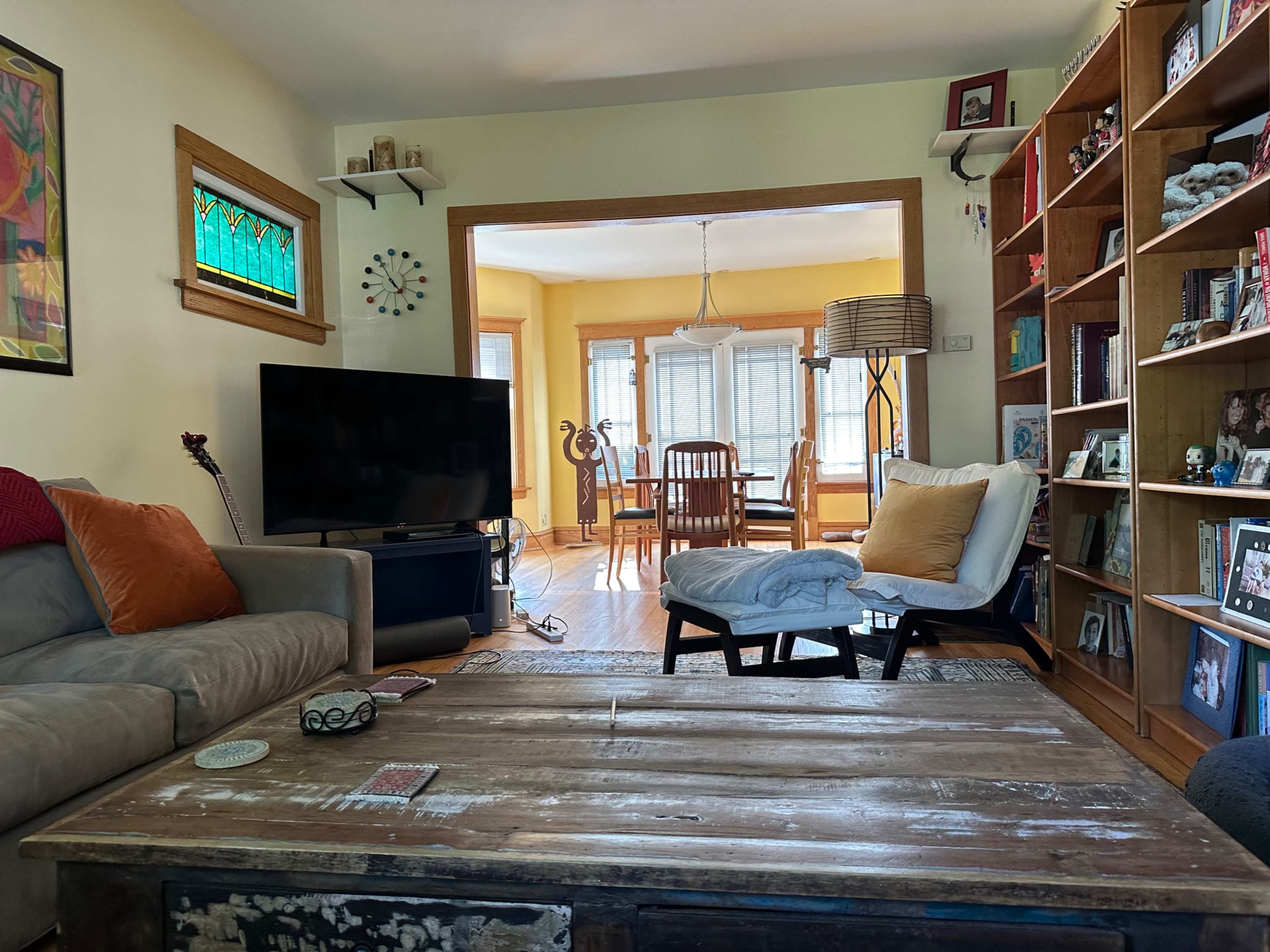 A cozy living room with a sofa, coffee table, and a television, with a dining area visible in the background featuring a light fixture and large windows.