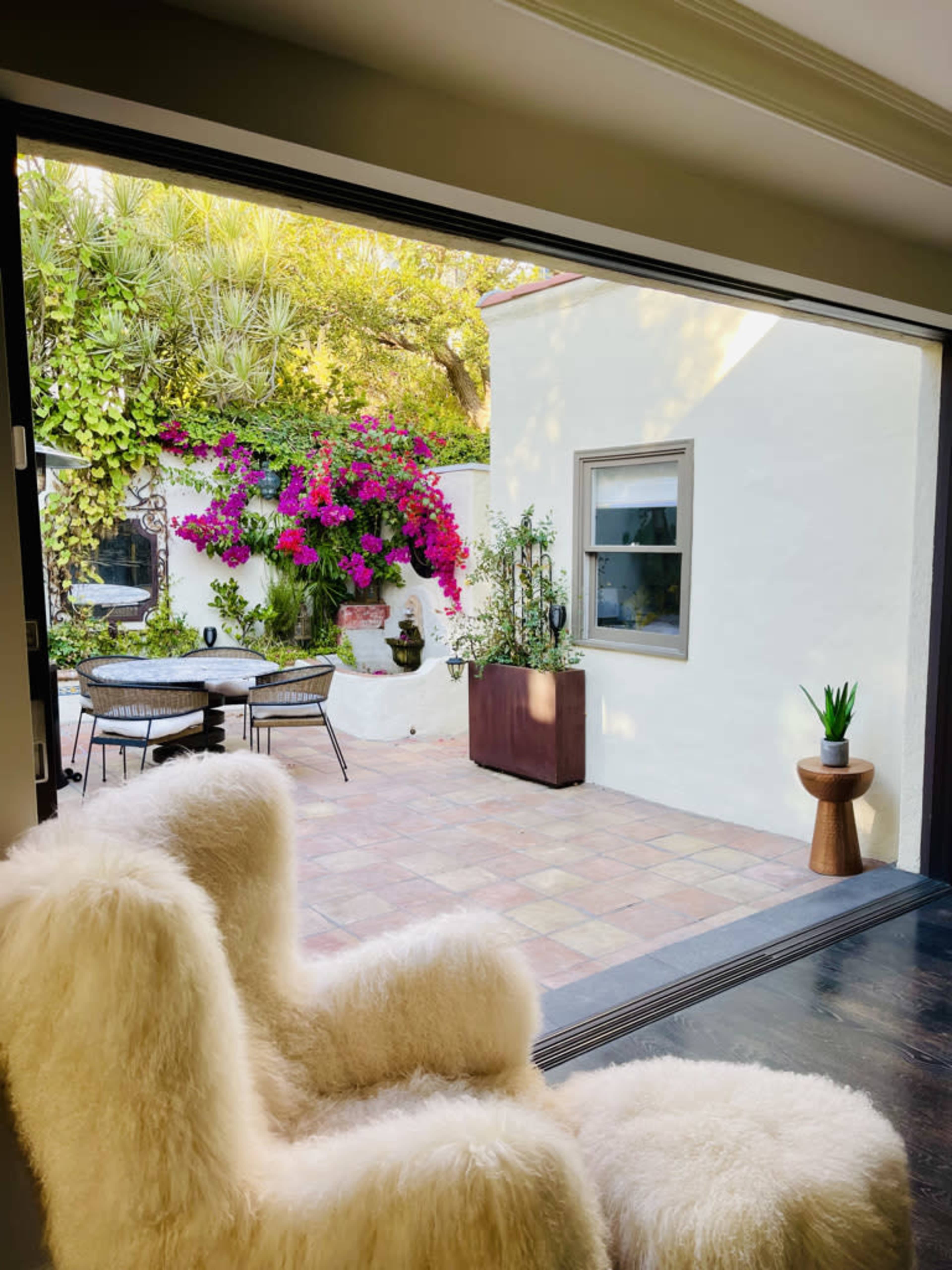 A view from a room features a patio with a round table and chairs, surrounded by lush greenery and vibrant bougainvillea.