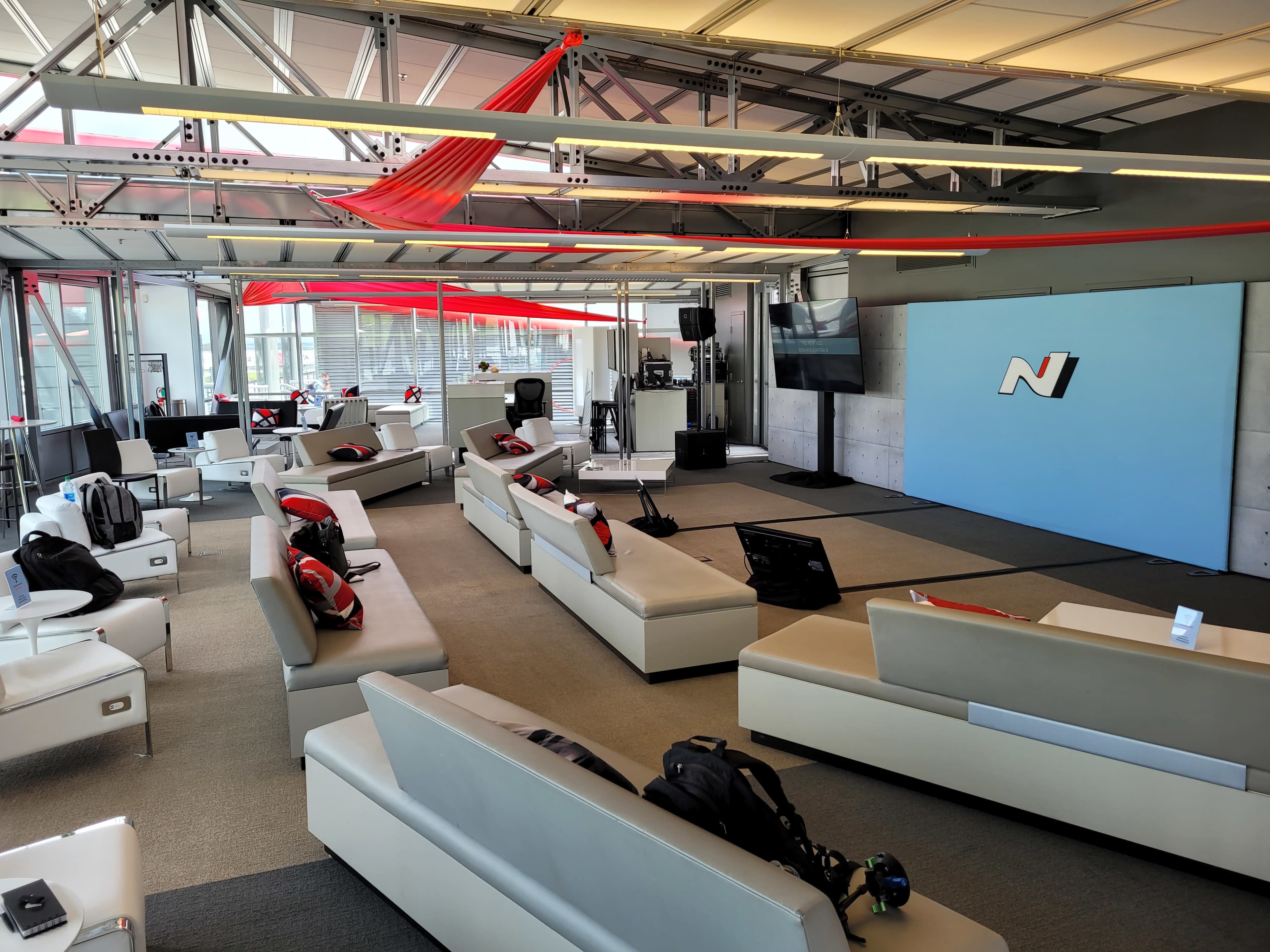 A modern lounge area with white sofas, a television screen, and red accents in a spacious setting.