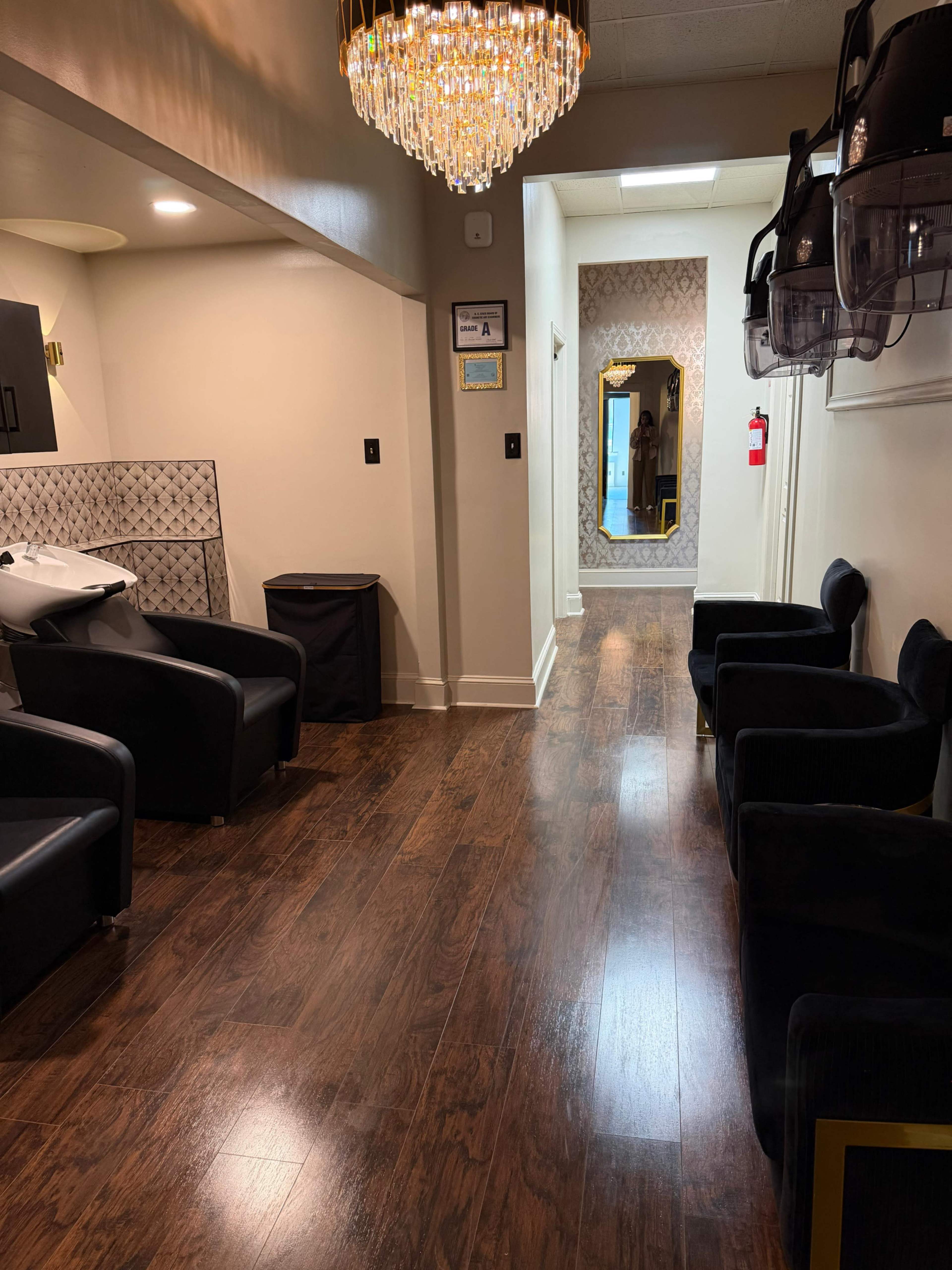 The image shows a well-lit salon interior featuring black seating, a chandelier, and mirrored walls, with a hallway leading to additional rooms.