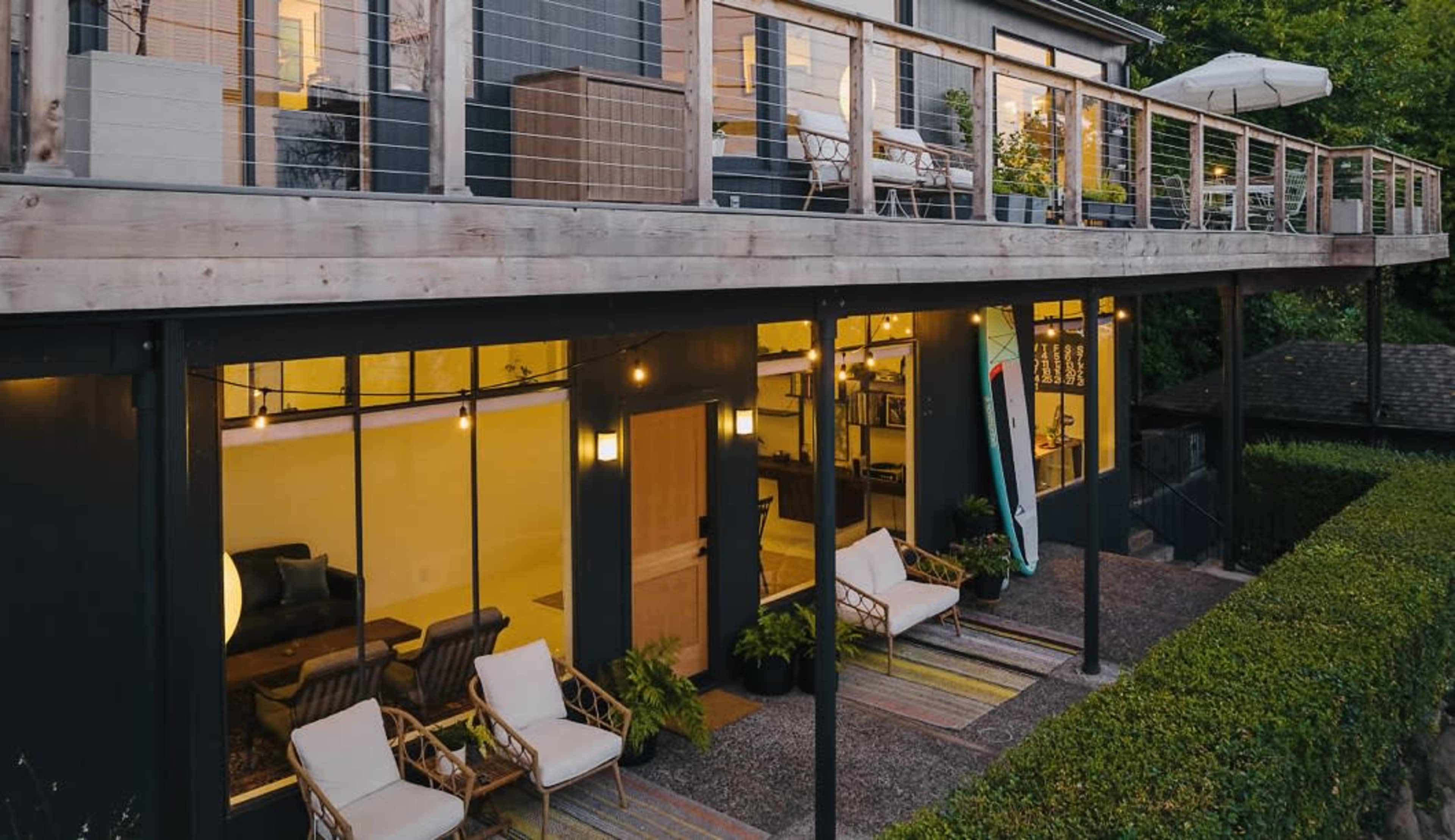 A two-story building with a wooden deck, outdoor seating, and large glass windows, surrounded by greenery.