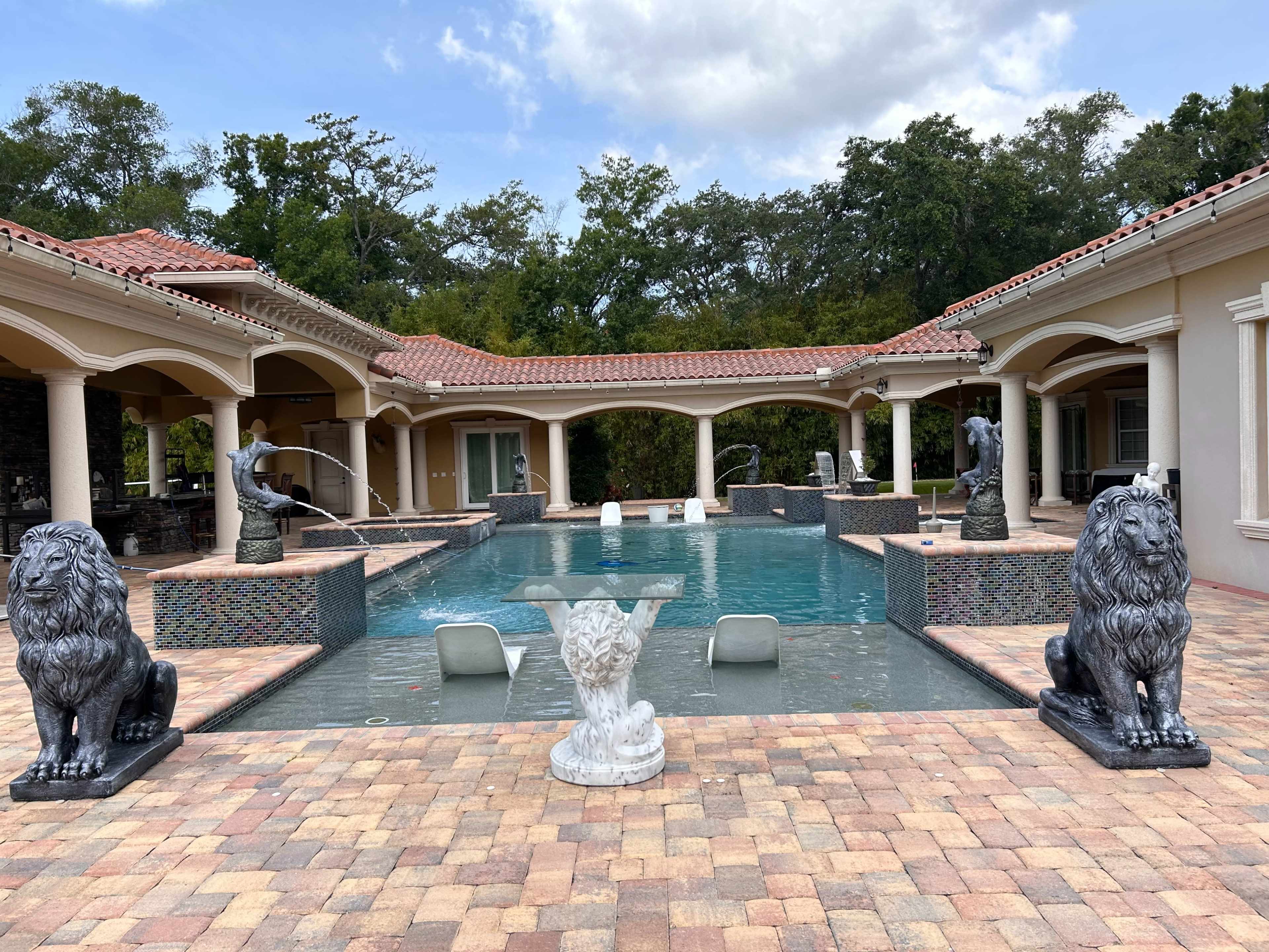 A large swimming pool surrounded by a tiled patio, featuring two lion statues at each end and sculptures on either side.