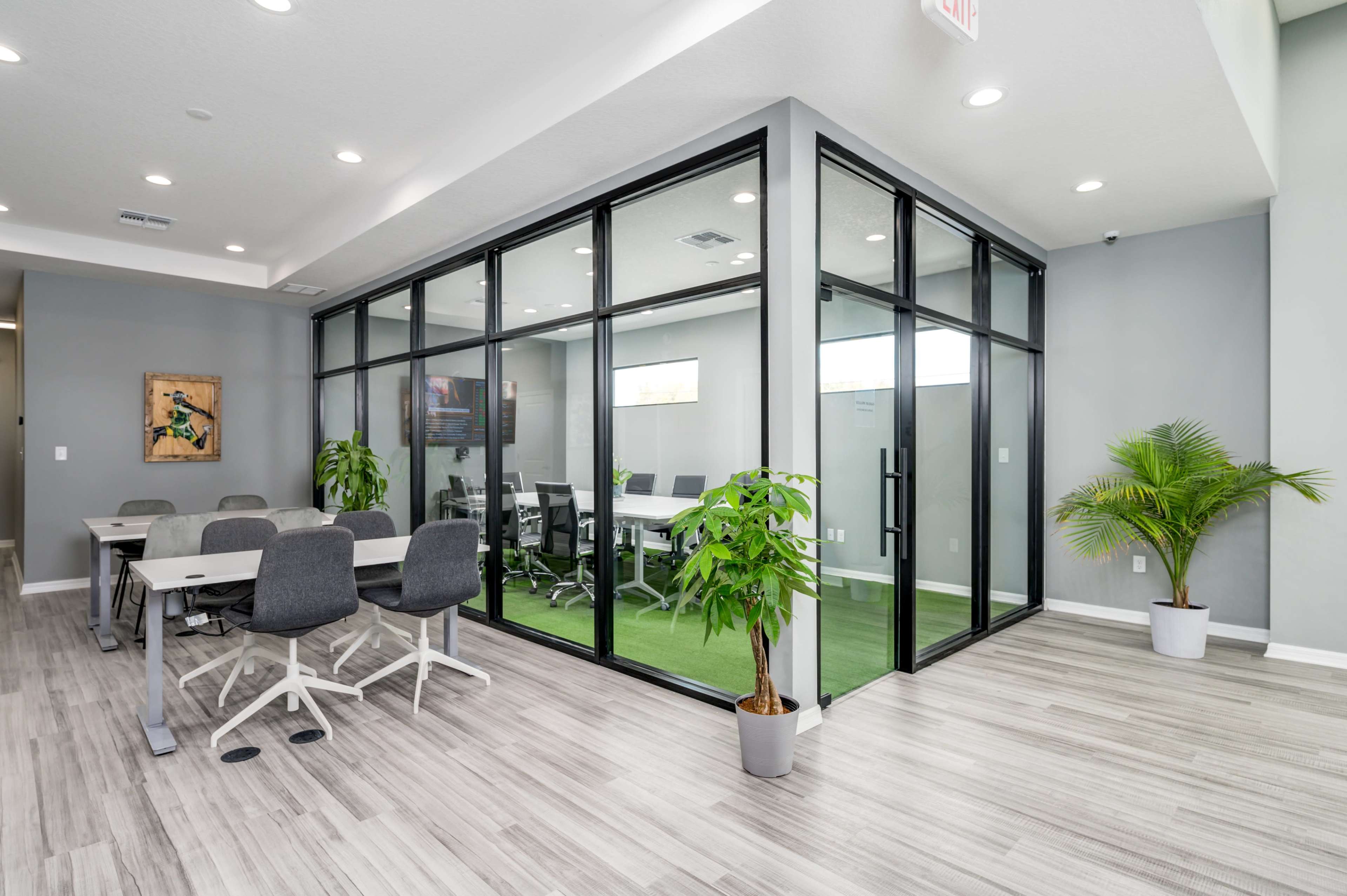 The image shows a modern office space with a glass-walled meeting room, desks arranged in a collaborative area, and potted plants for decor.