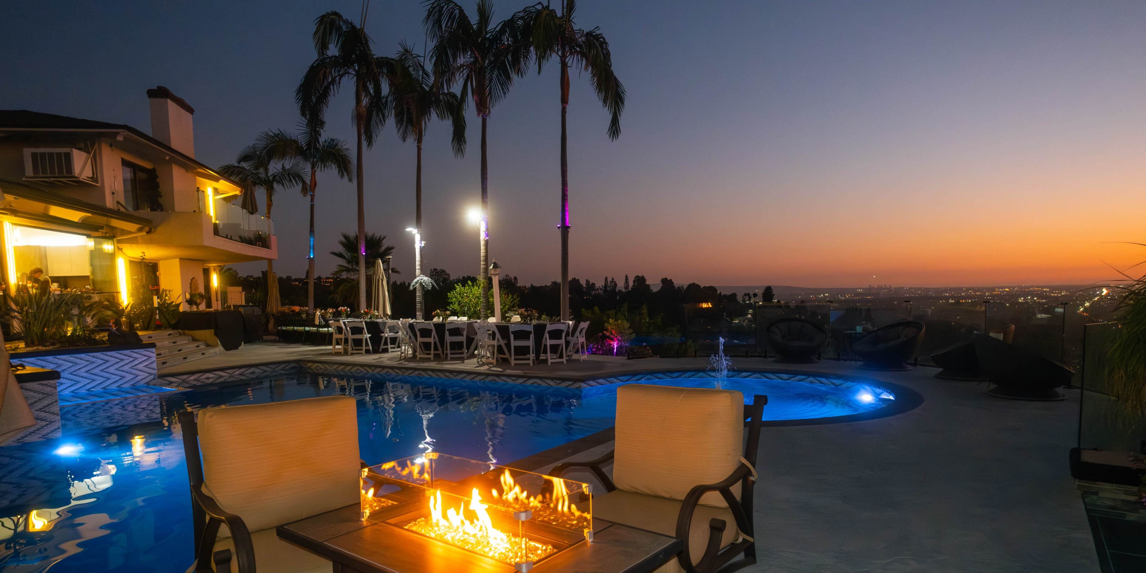 A luxurious outdoor space features a pool with a fire table, surrounded by palm trees and illuminated lounge chairs, set against a sunset view.