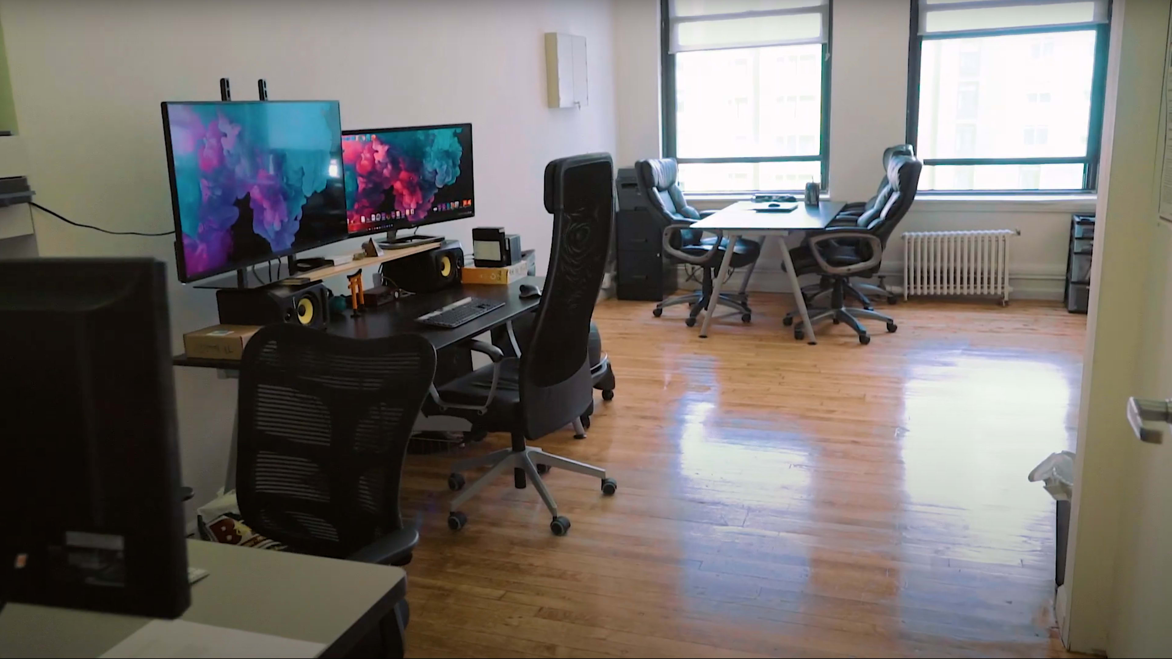 A modern office space with two workstations featuring computer monitors and ergonomic chairs, empty desks, and large windows allowing natural light to enter.