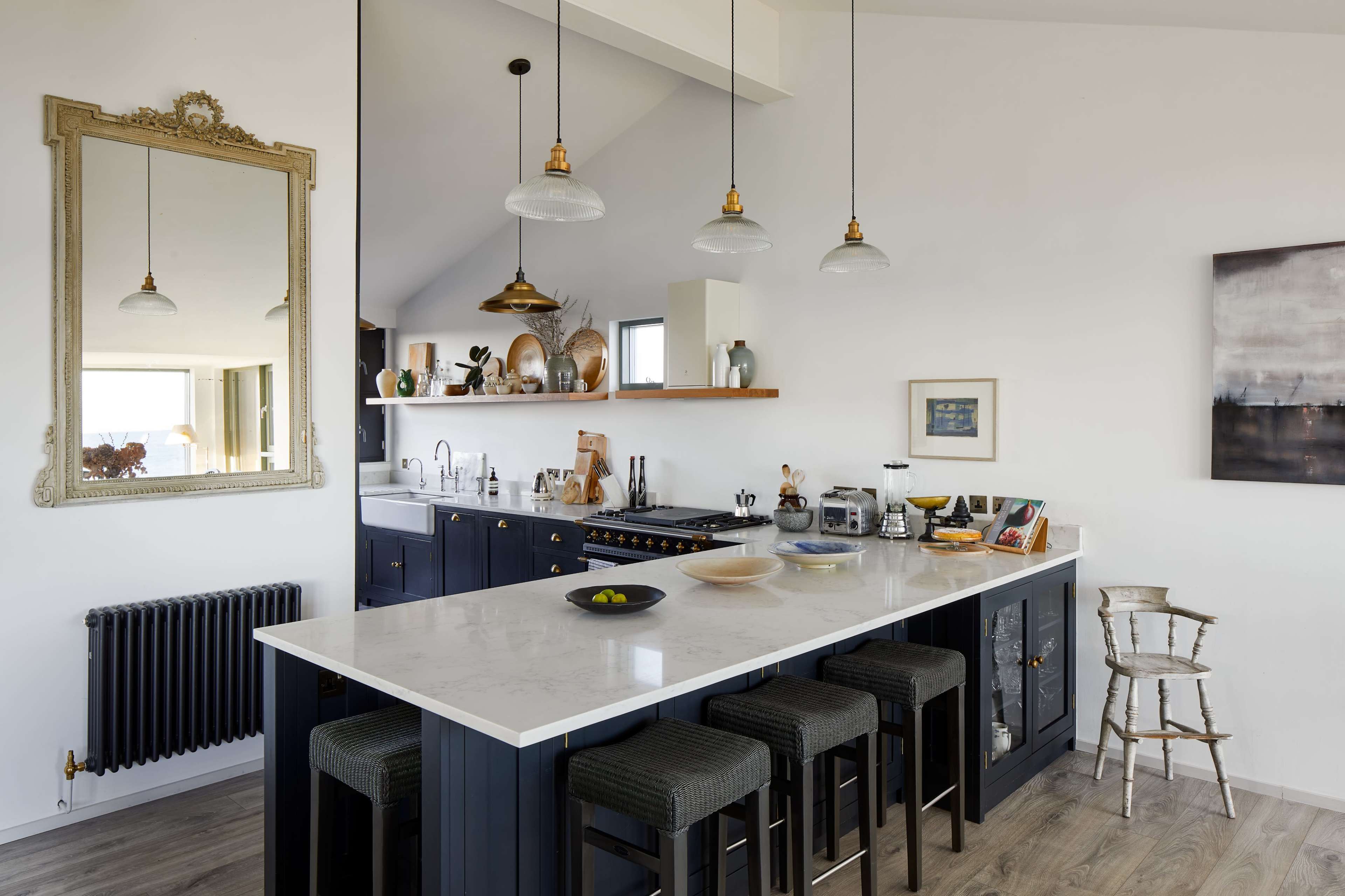 The image depicts a modern kitchen with a large island, four stools, a marble countertop, and a mix of dark cabinetry and wooden shelves, along with a mirror and pendant lights.