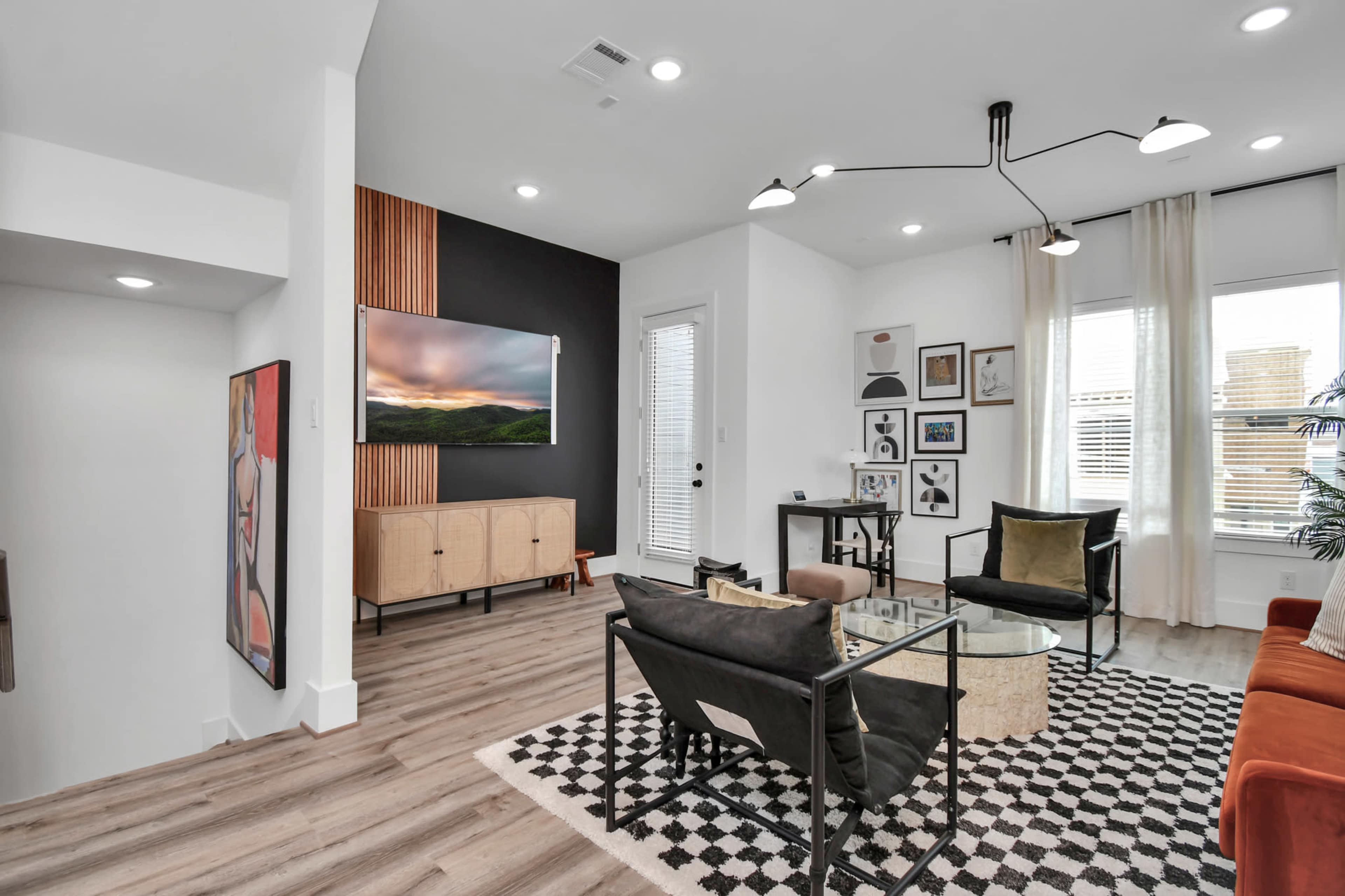 The image shows a modern living room with a television mounted on a wall, a glass coffee table, and a mix of seating options against a backdrop of various artwork and plants.