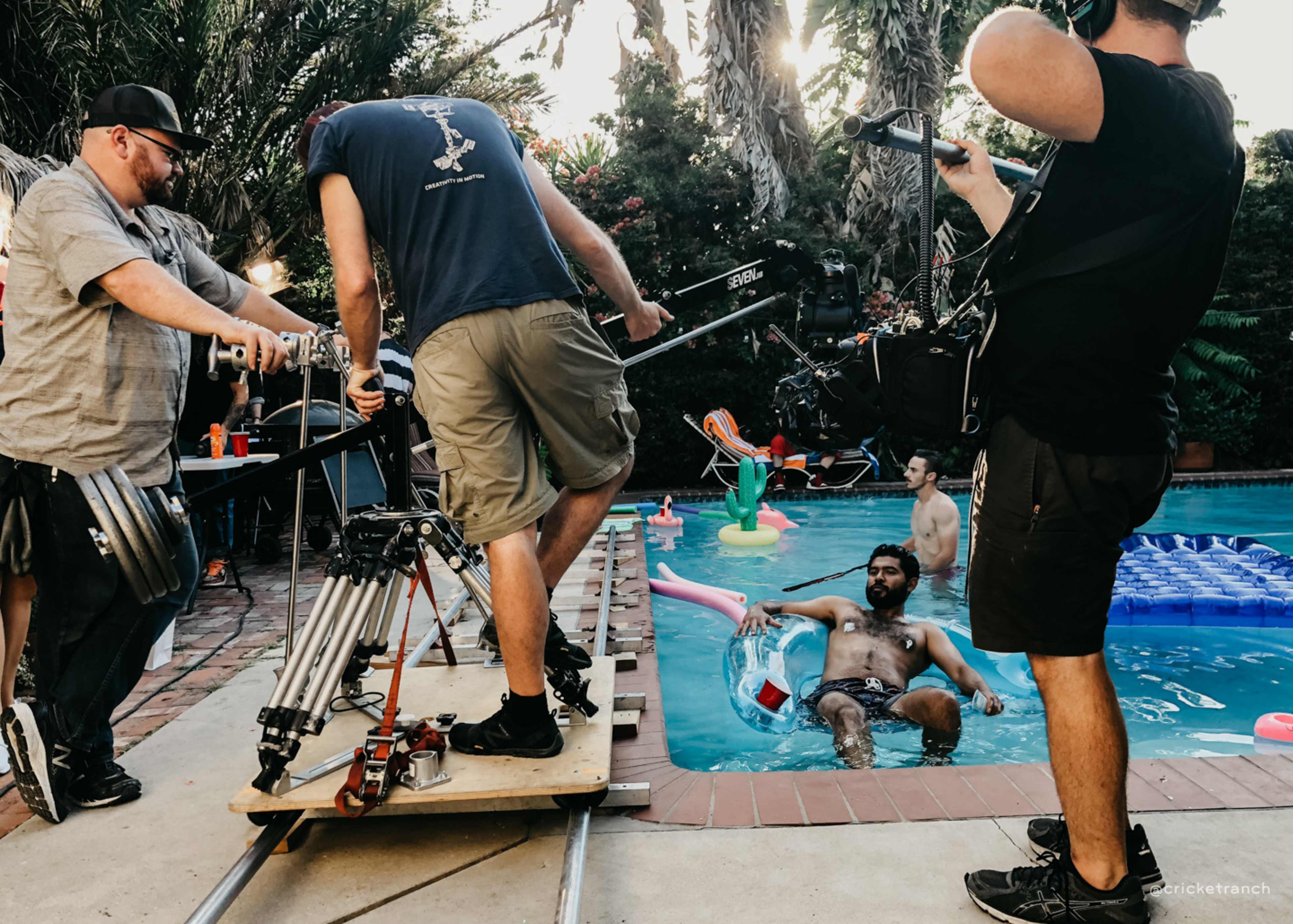 A film crew is set up by a swimming pool, with one person sitting on an inflatable float while another operates a camera on a dolly.