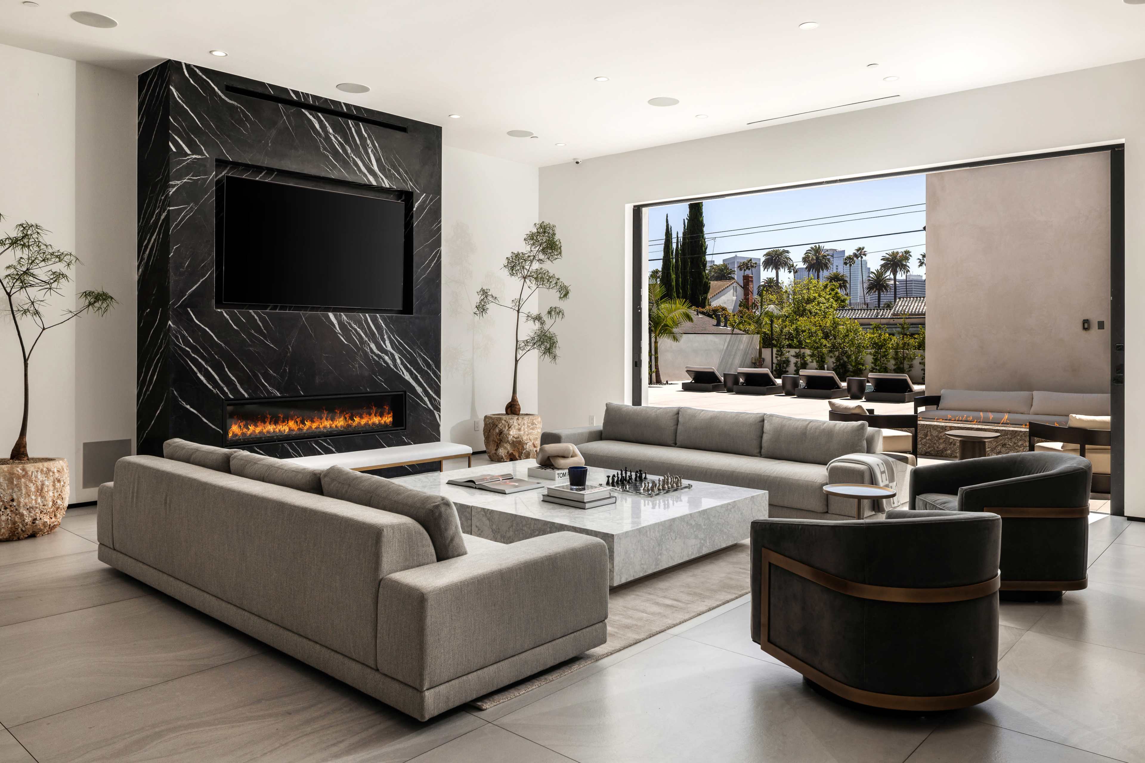 The image shows a modern living room featuring a large black marble fireplace with a TV mounted above it, sleek gray sofas, and a glass wall that opens to an outdoor patio area.