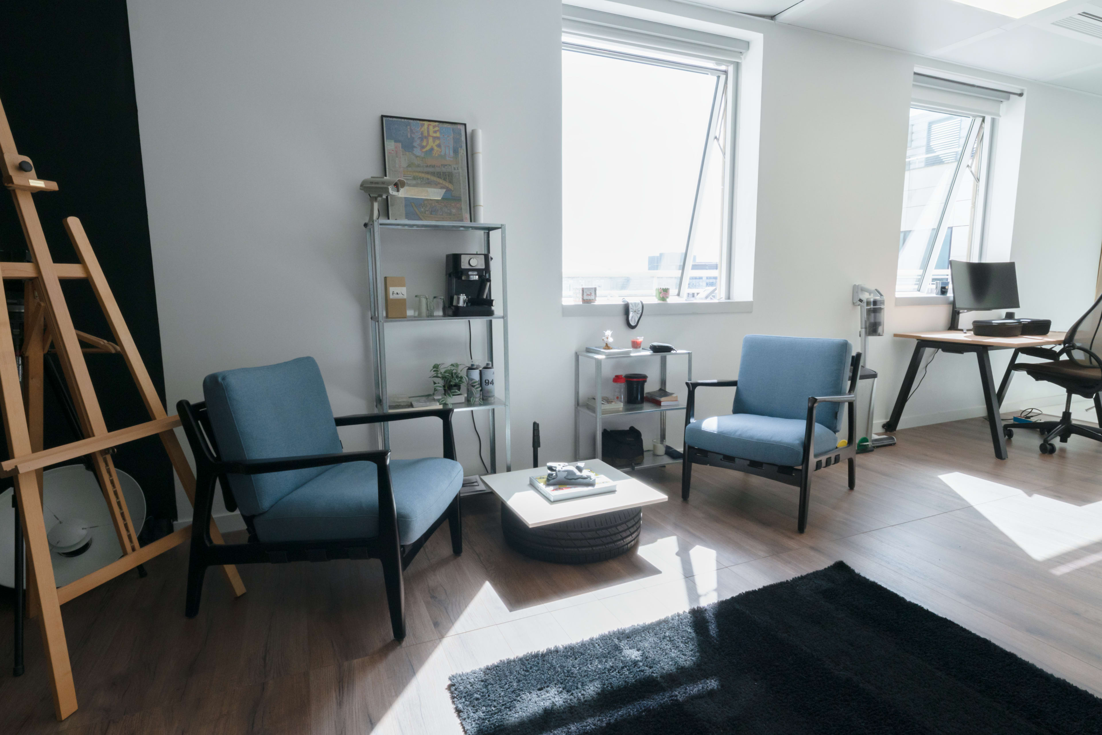 The image shows a modern office space with two blue chairs, a small coffee table, and an easel, illuminated by sunlight through large windows.