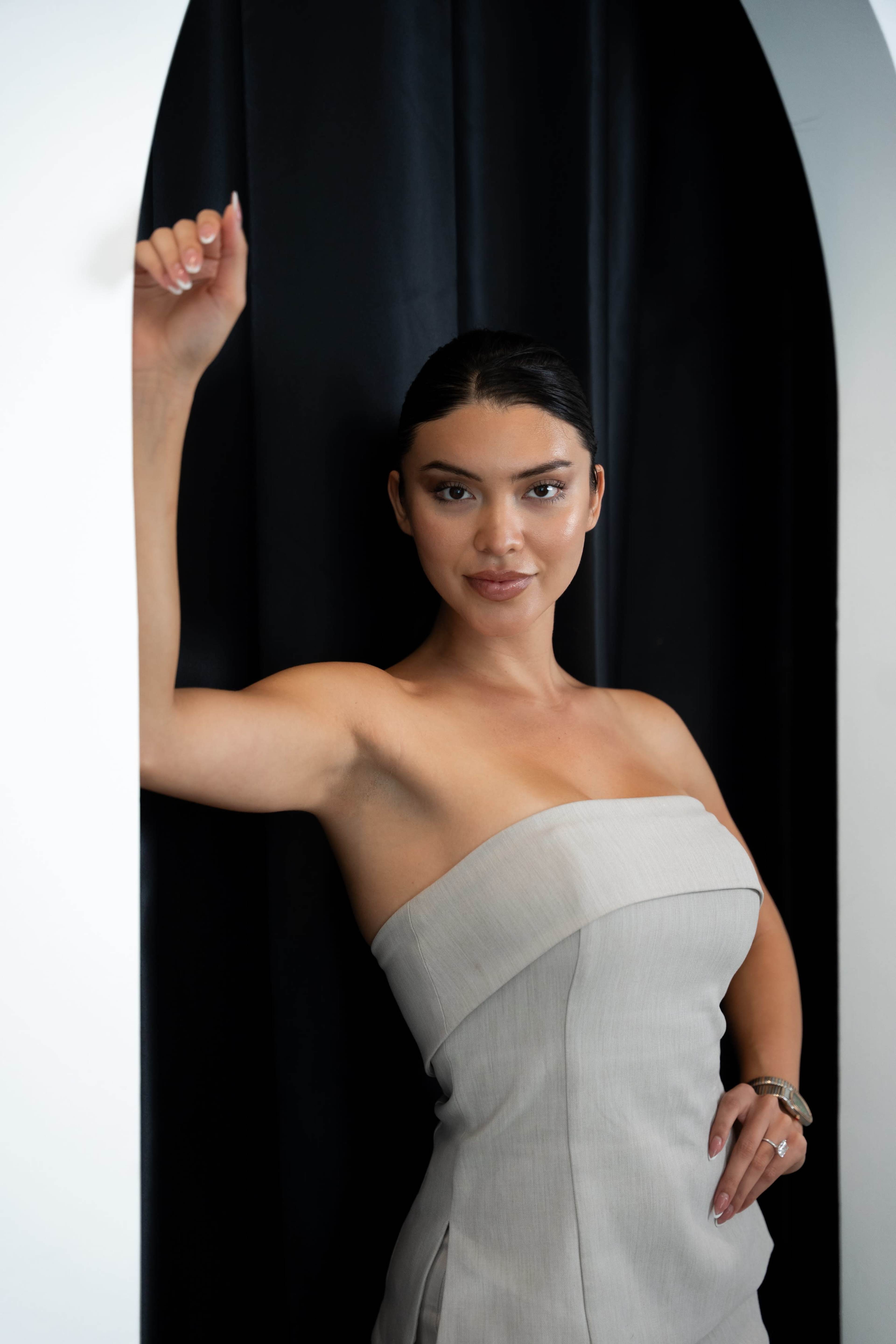 A woman in a strapless beige dress poses confidently in a doorway with black curtains.