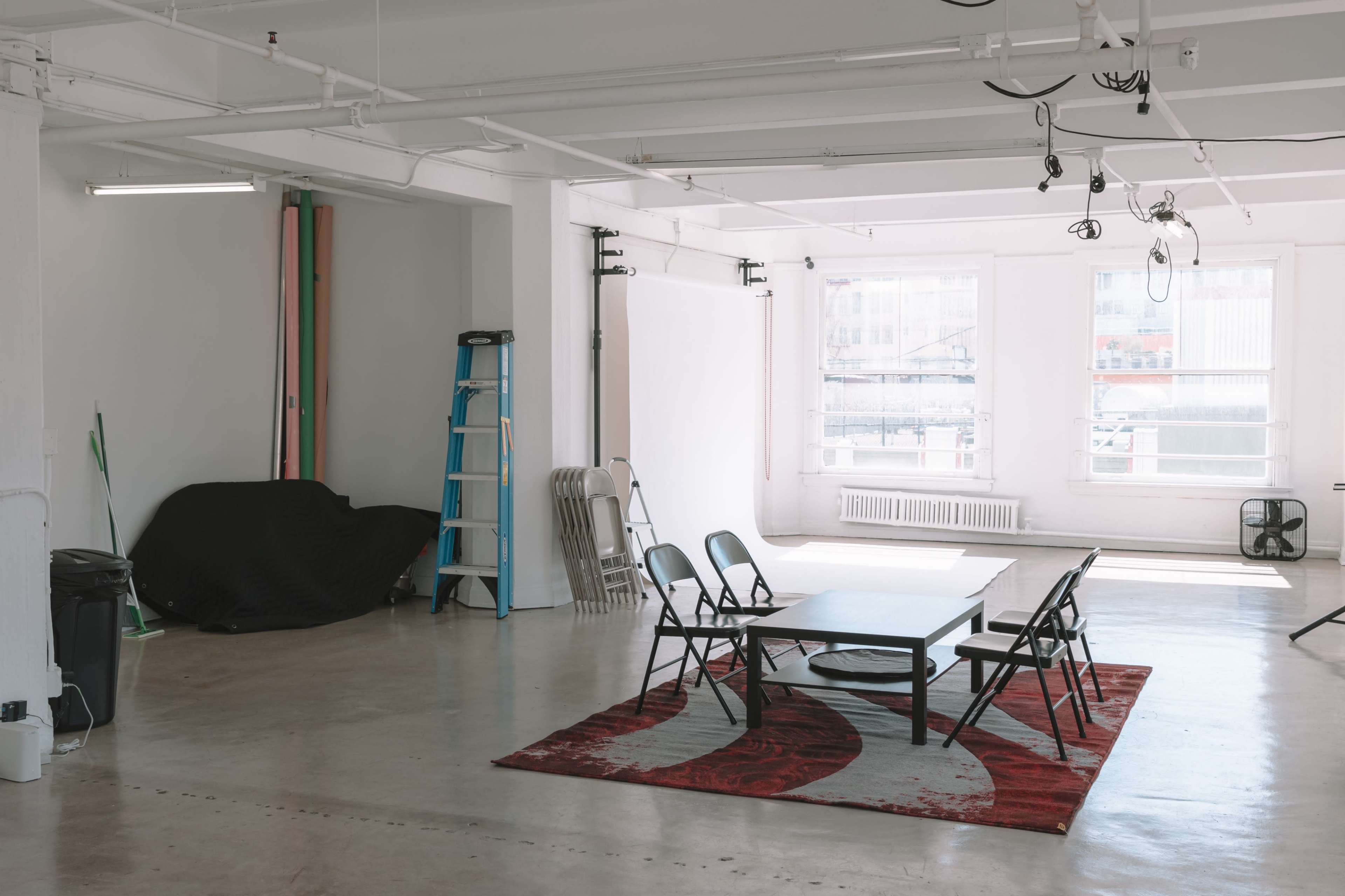 An empty studio space features a table surrounded by black chairs, a large carpet, and various equipment along the walls.