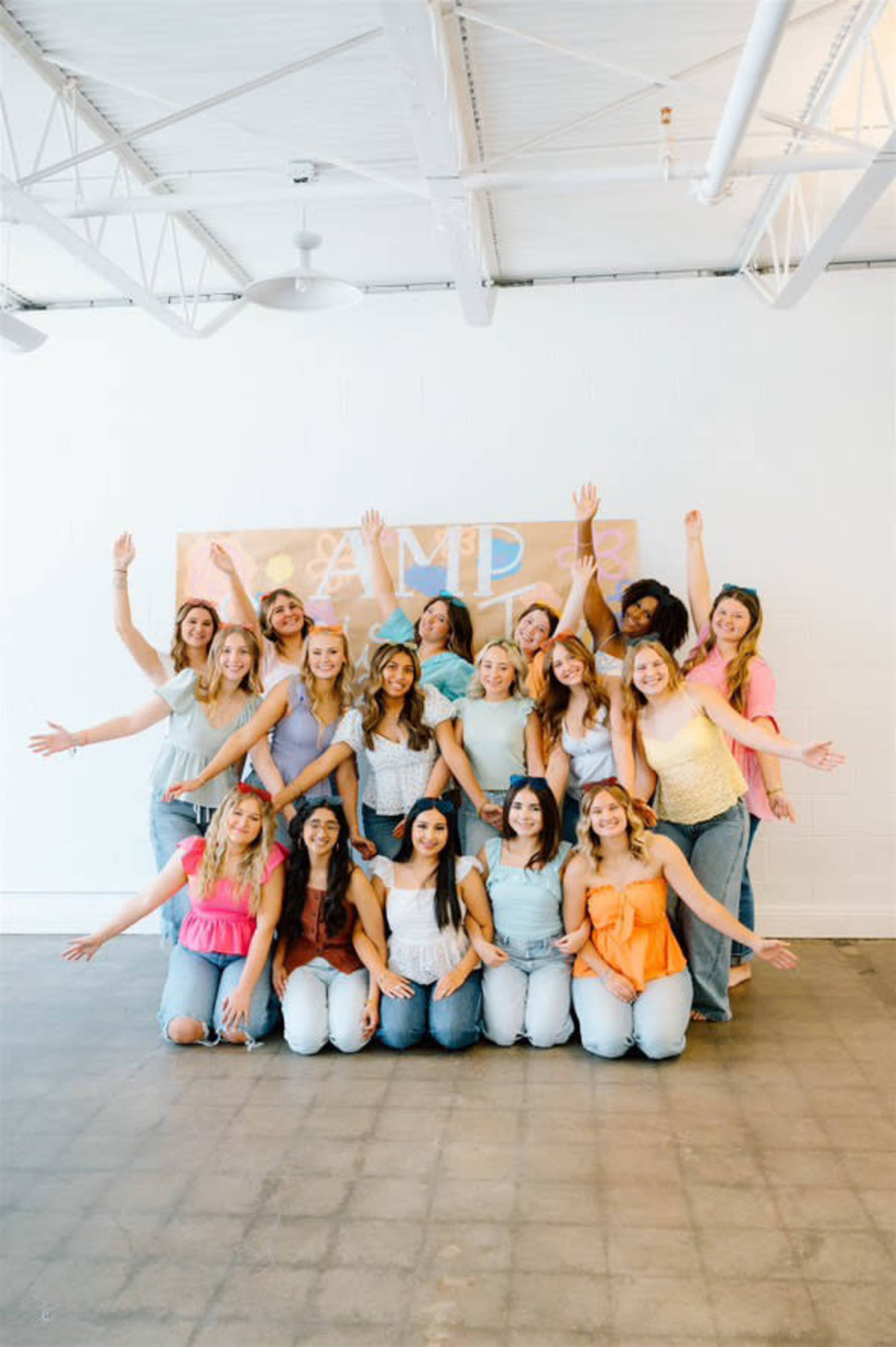 A large group of smiling women poses together with raised arms in a bright indoor space decorated with a colorful banner.