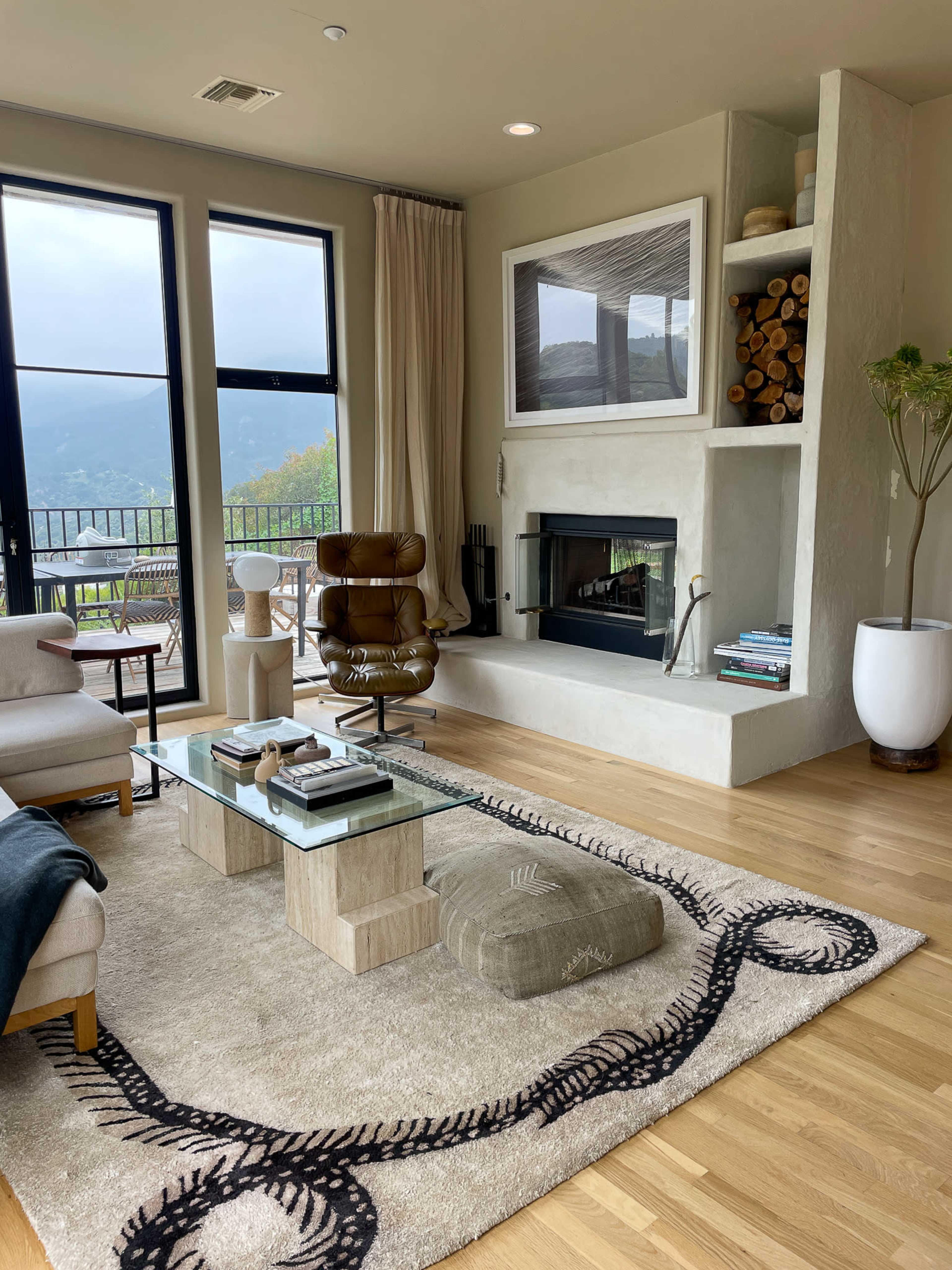The living room features a fireplace with stacked logs, a glass coffee table, a comfortable chair, and large windows overlooking a mountainous view.