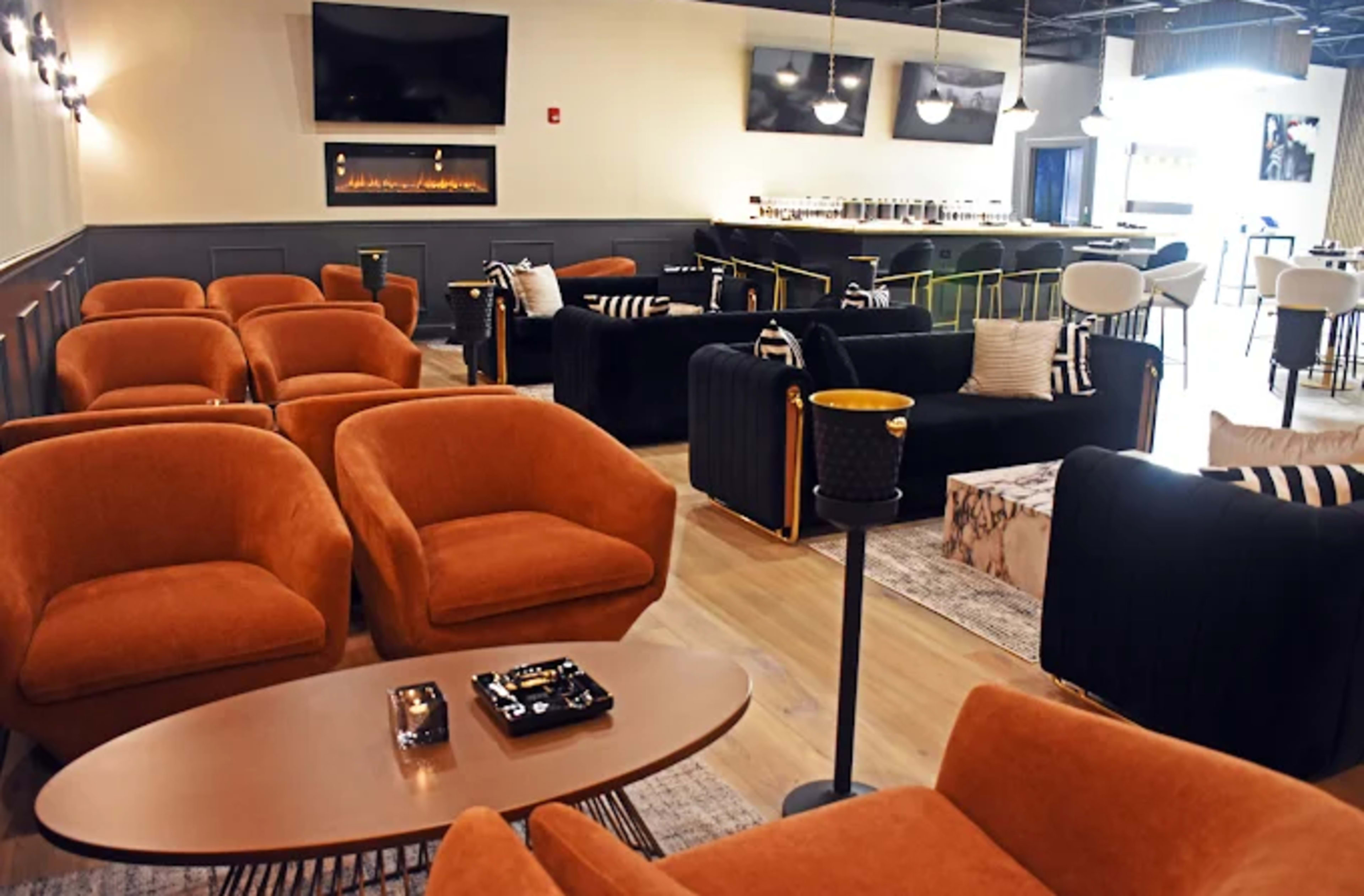 The image shows a modern lounge area featuring orange upholstered chairs, a low table, and a stylish bar with seating and decorative lighting.