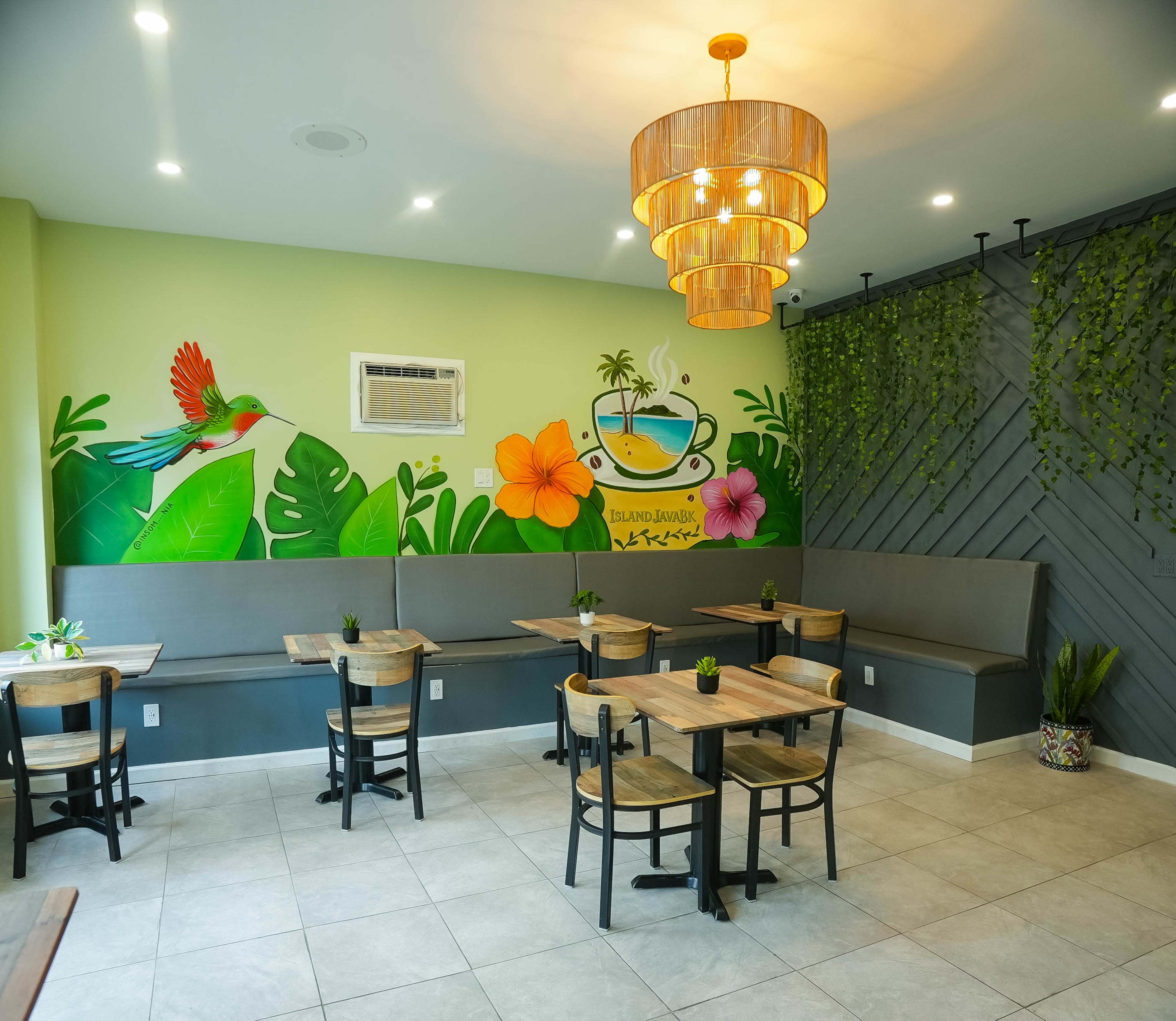 The image shows a brightly colored café interior featuring a mural of a tropical scene, several wooden tables and chairs, and a decorative light fixture.
